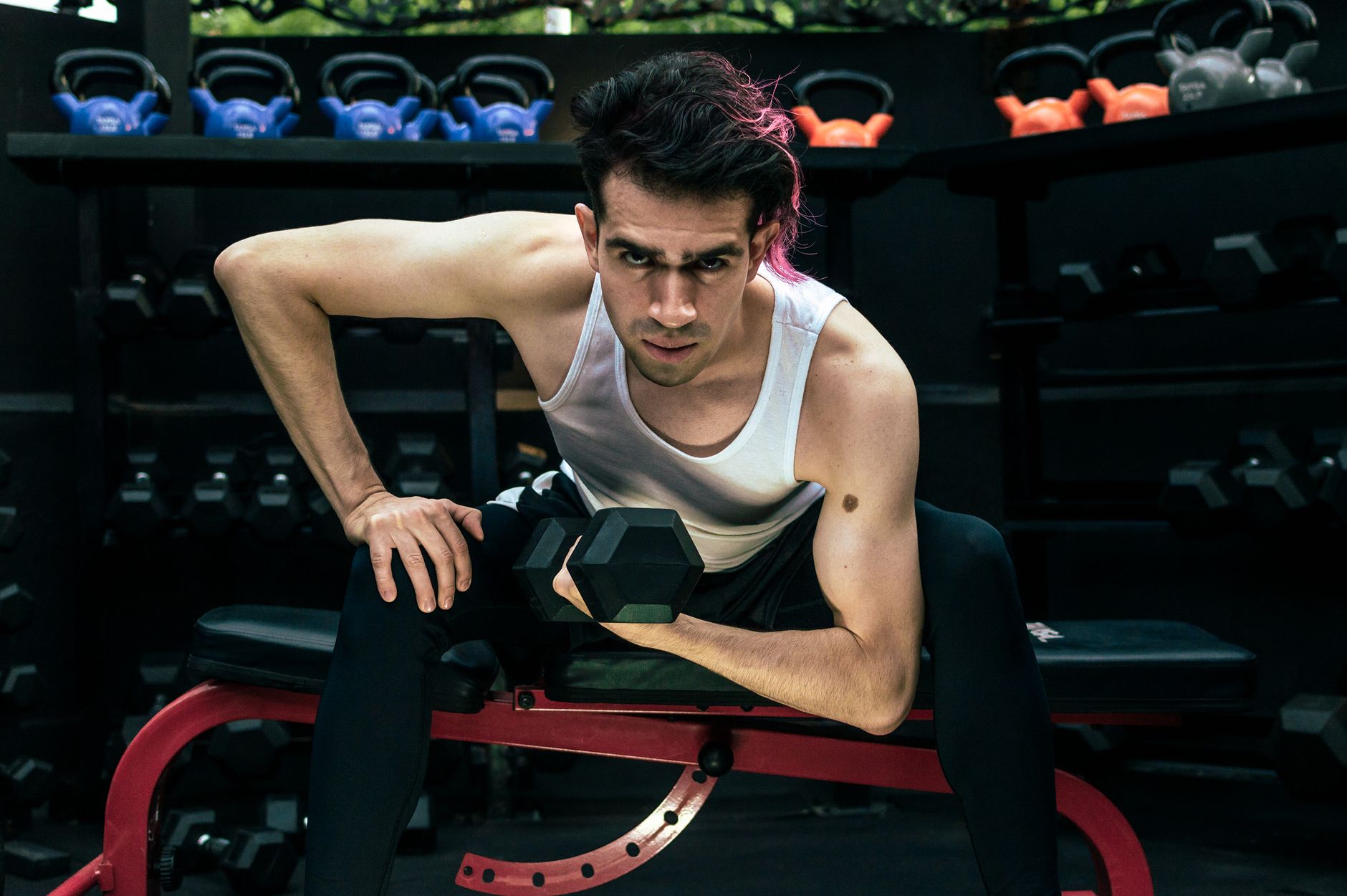 A man intensely lifting a dumbbell on a bench in a gym environment.