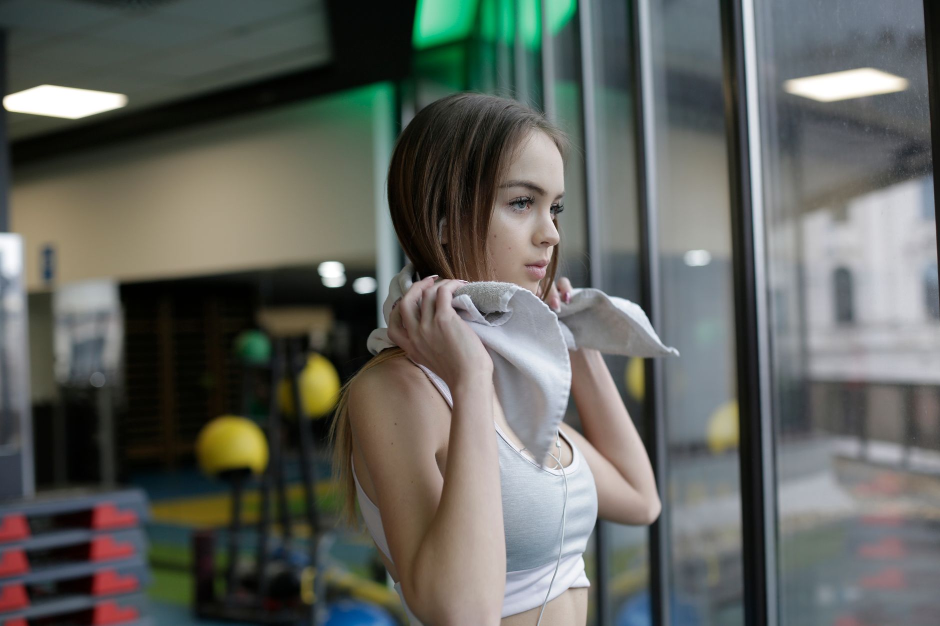 Serious young female athlete in white sportswear standing with towel around neck near window in modern gym and looking away