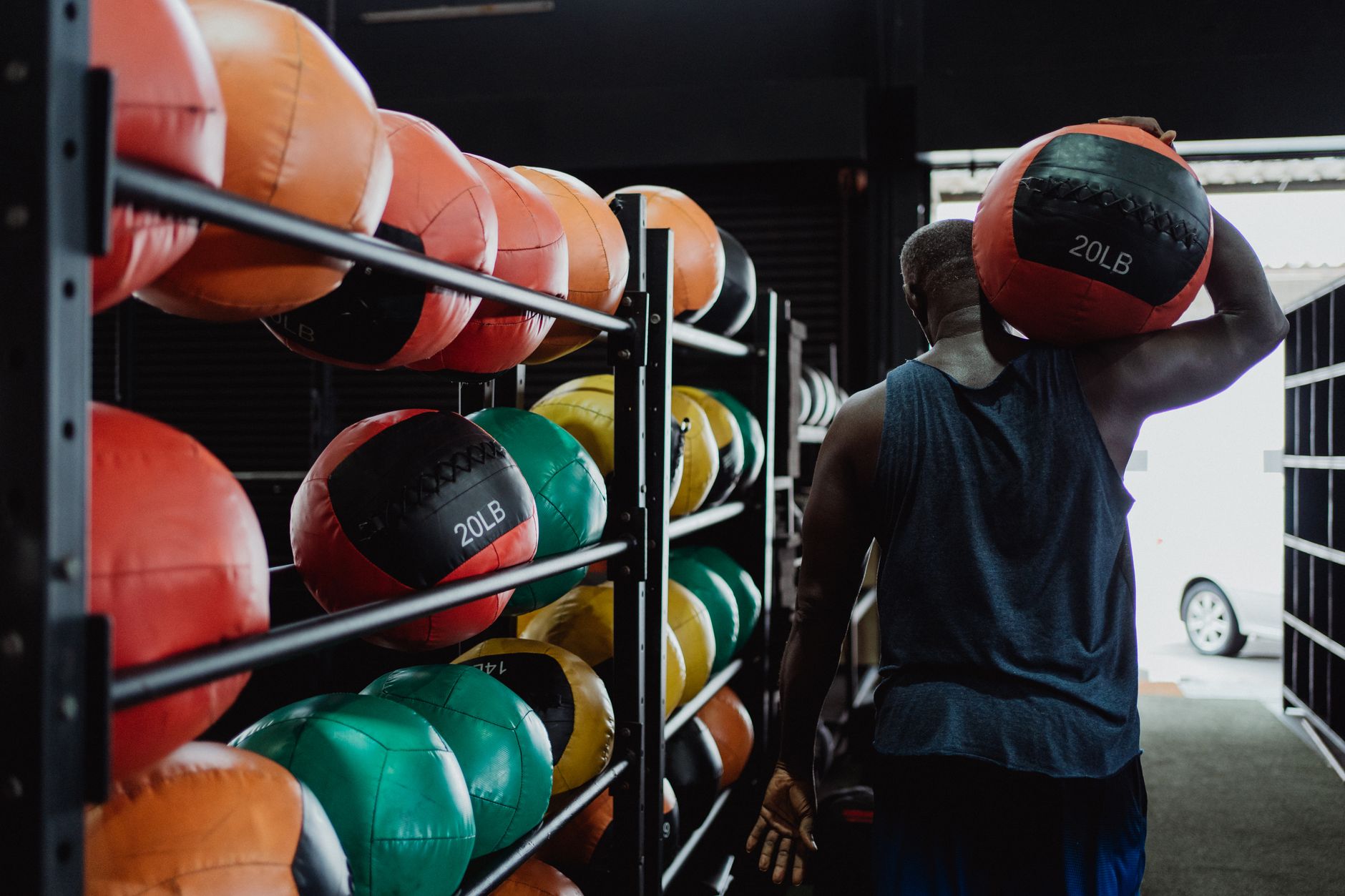 Back view of a man carrying a 20LB medicine ball in a gym with colorful balls on racks.