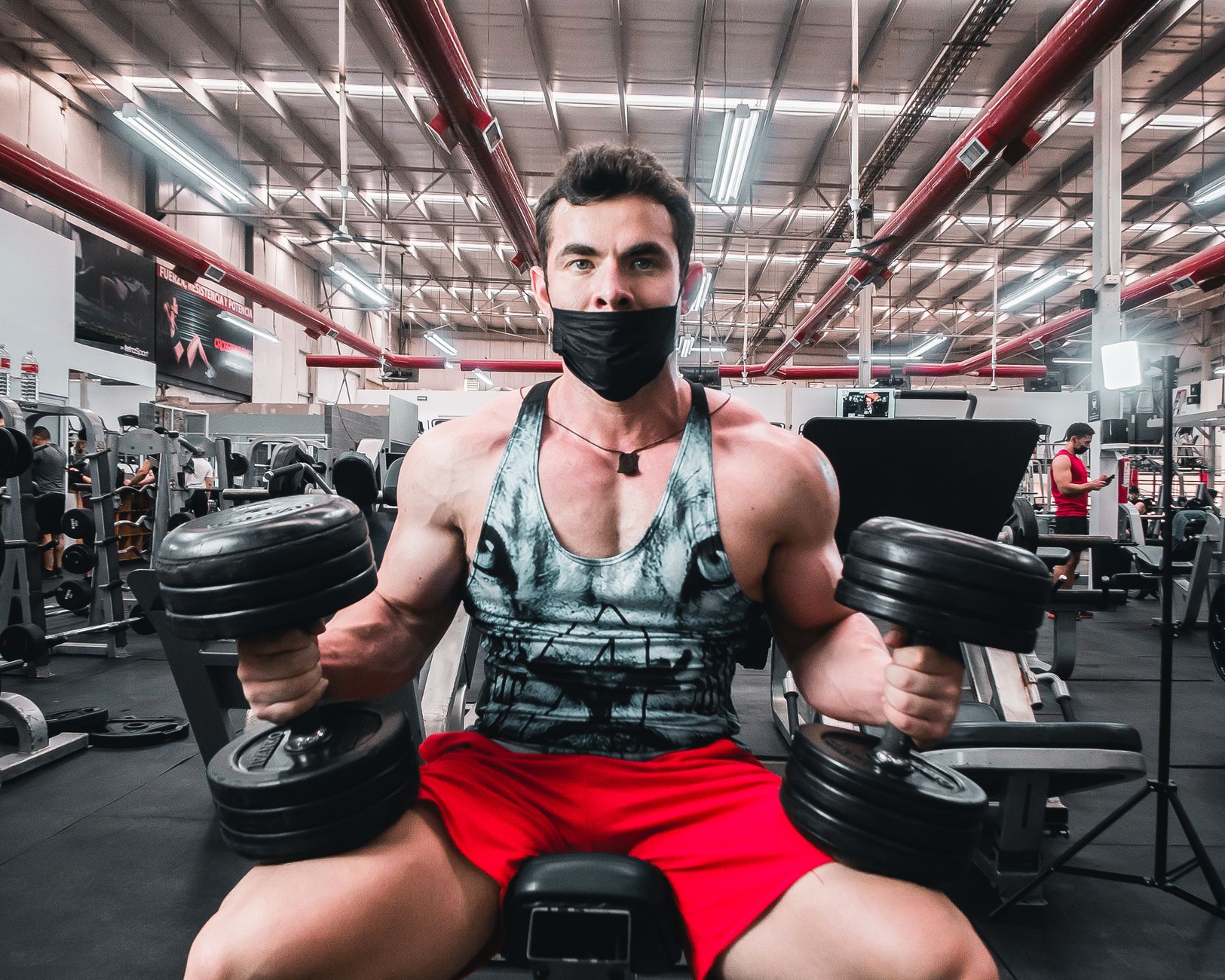 Muscular man lifting dumbbells while wearing a face mask in a modern gym setting.