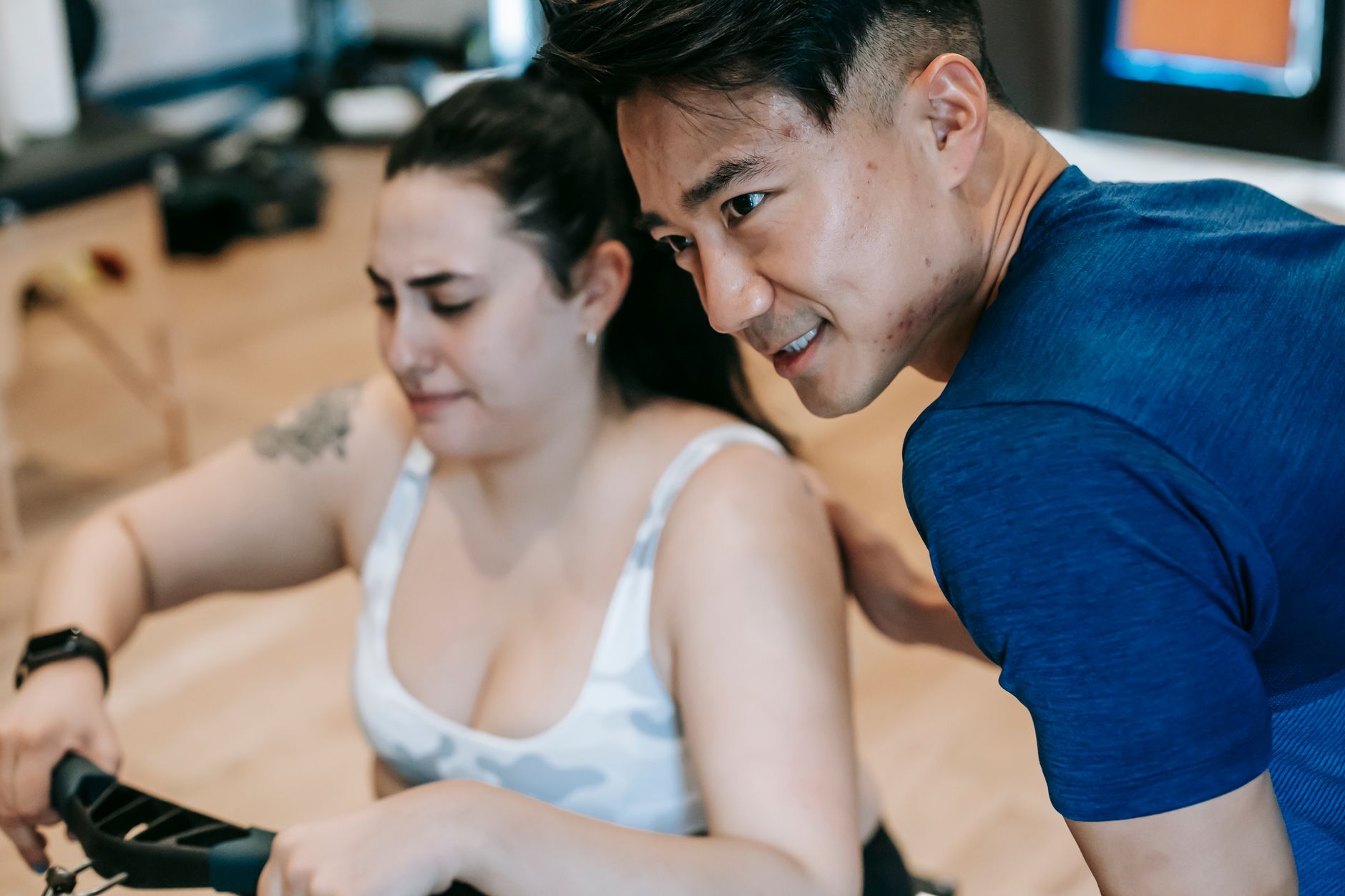 Side view of crop positive Asian male couch supporting young plus size female doing exercise on gym equipment during intense workout