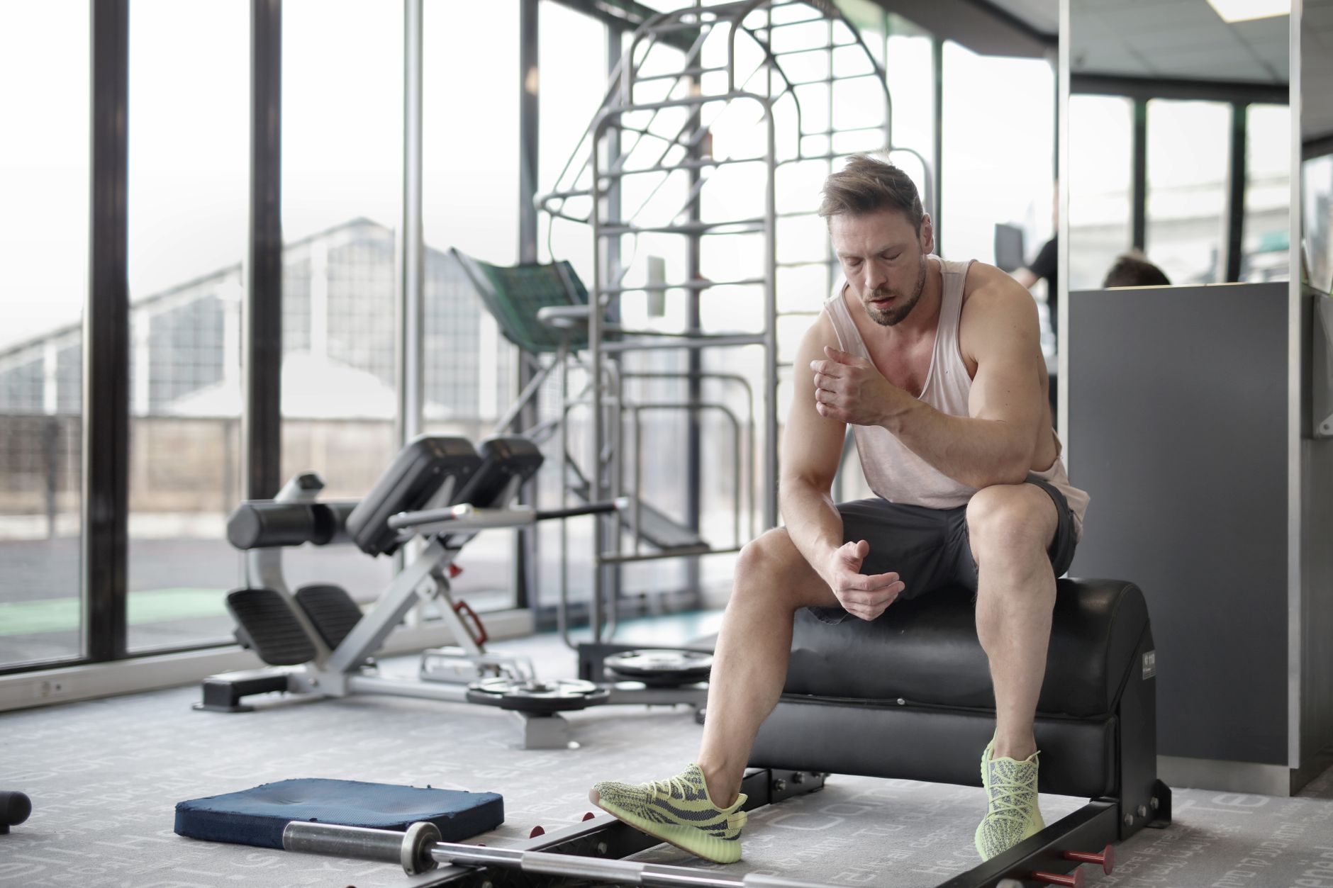 Tired young muscular male athlete in casual sportswear and sneakers sitting on bench and resting after intense weightlifting workout in modern fitness club