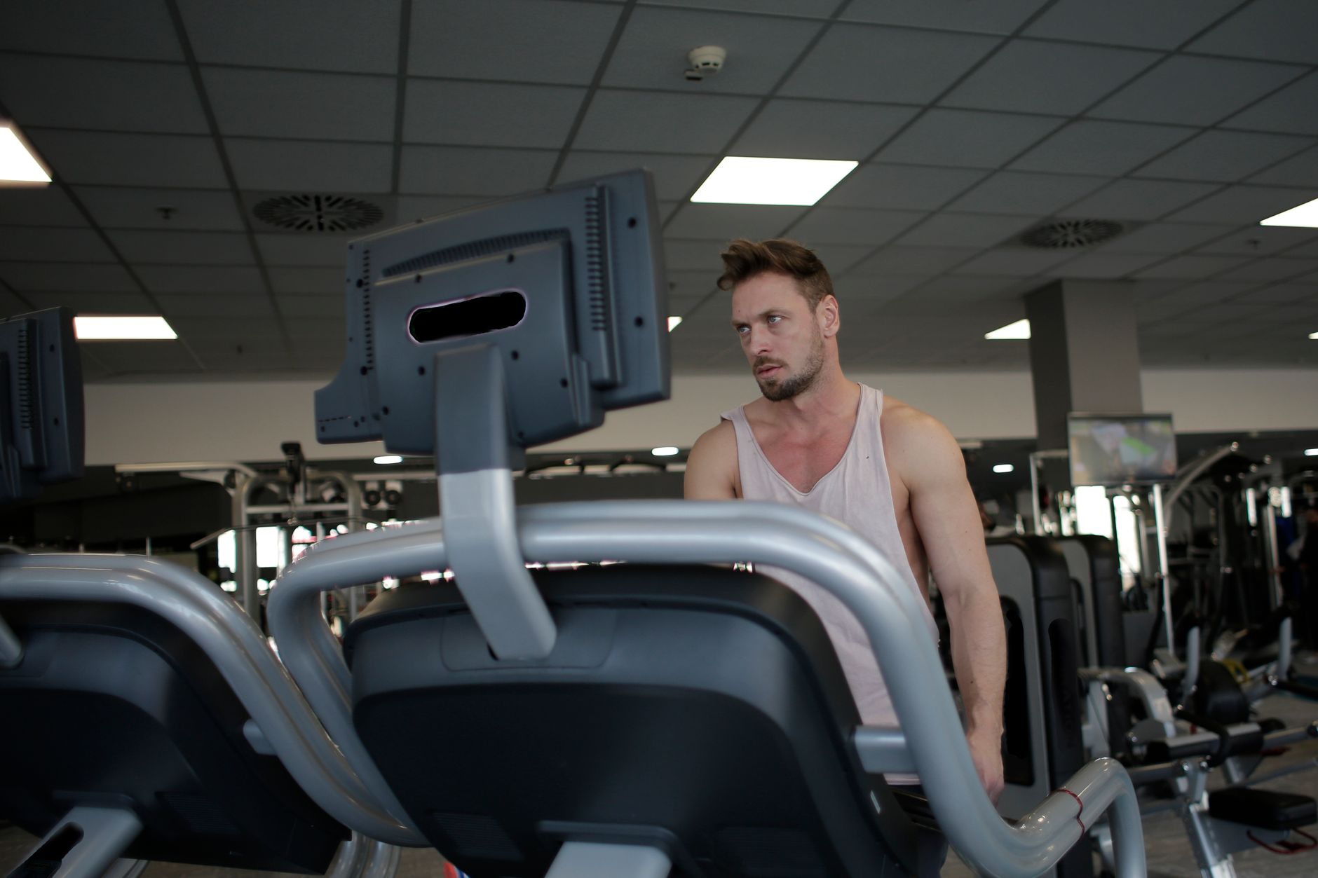 Confident muscular adult male athlete in sportswear standing near running machine in modern spacious fitness center before training while looking away pensively