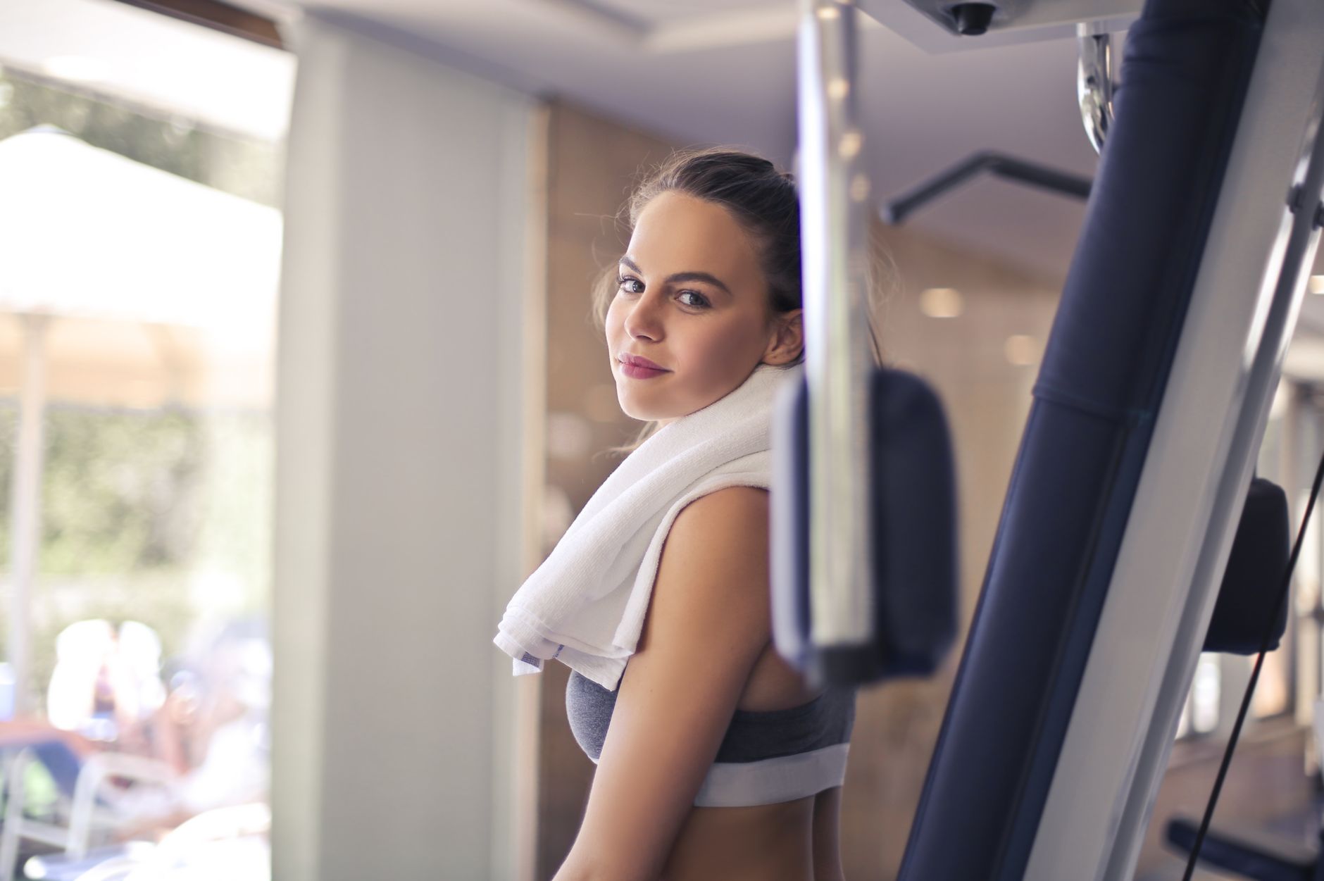 Low angle of slim female athlete in sportswear with towel on neck smiling at camera while standing beside sports equipment after training against blurred interior of light contemporary gym