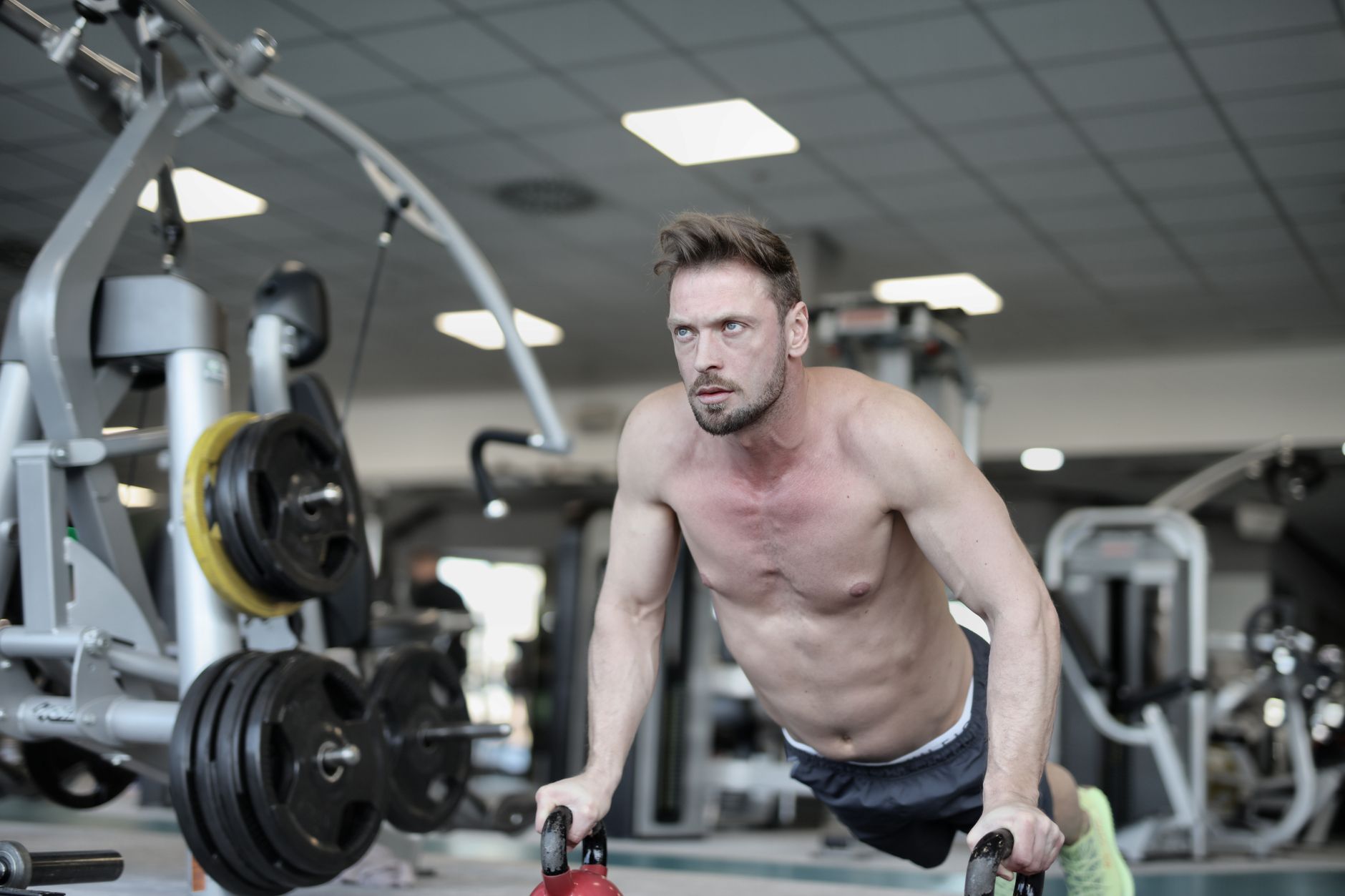Concentrate muscular sportsman with naked torso performing push ups on kettlebells and looking away while training in modern fitness club