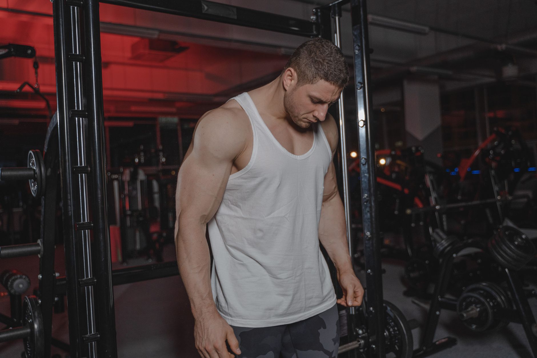 A muscular man in a gym wearing a white tank top, surrounded by weightlifting equipment.