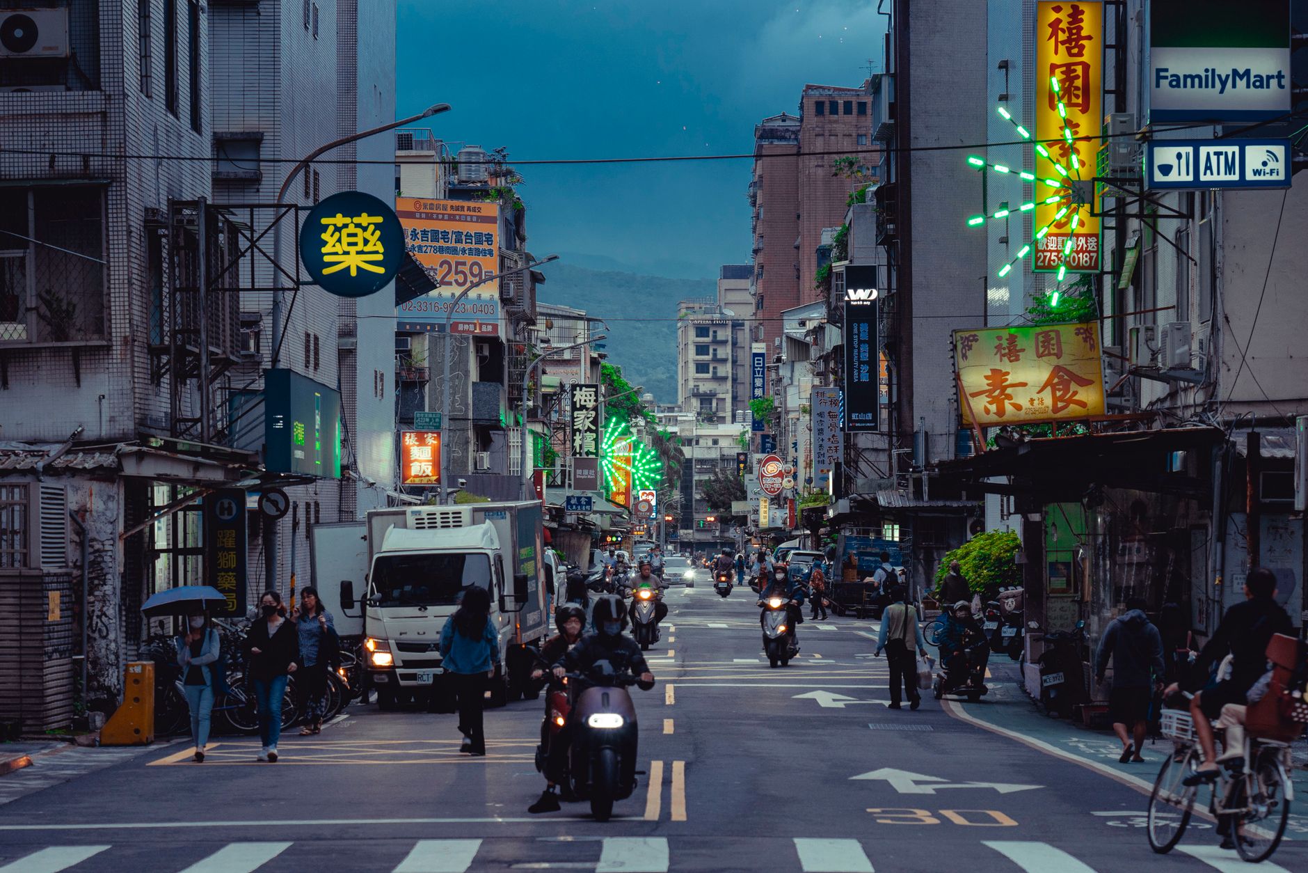 A lively city street scene with neon signs, scooters, and people in the evening.
