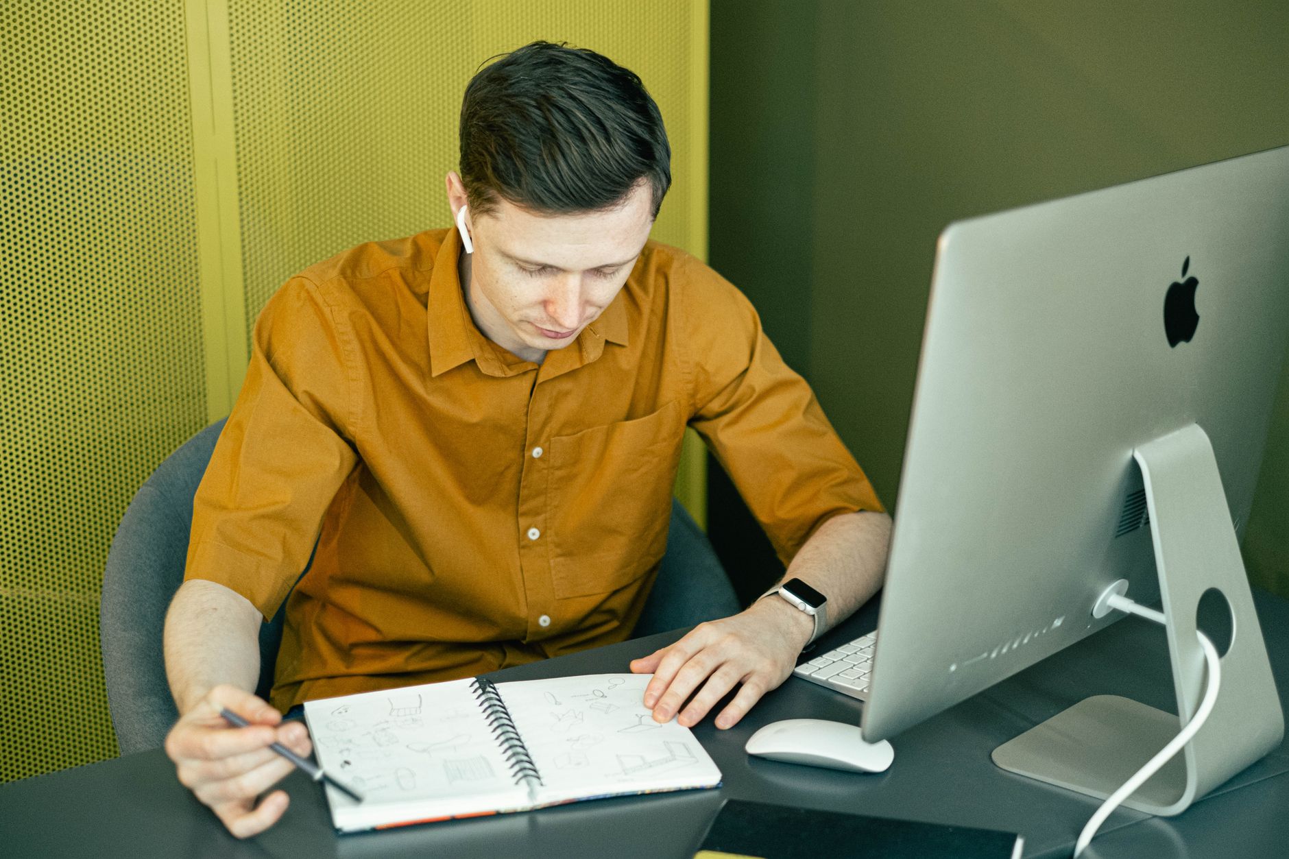 Focused young man sketching at his desk with a computer and notebook in a creative office setting.