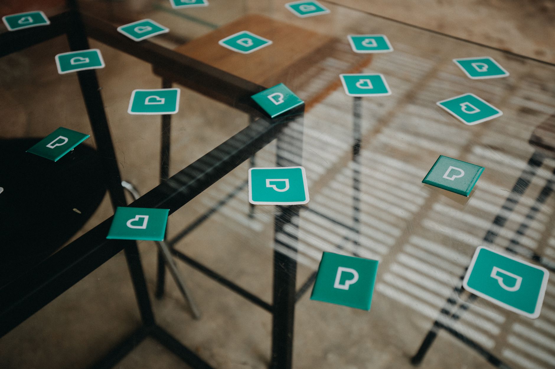 Scattered green logo magnets on a glass table seen from above, casting shadows in sunlight.