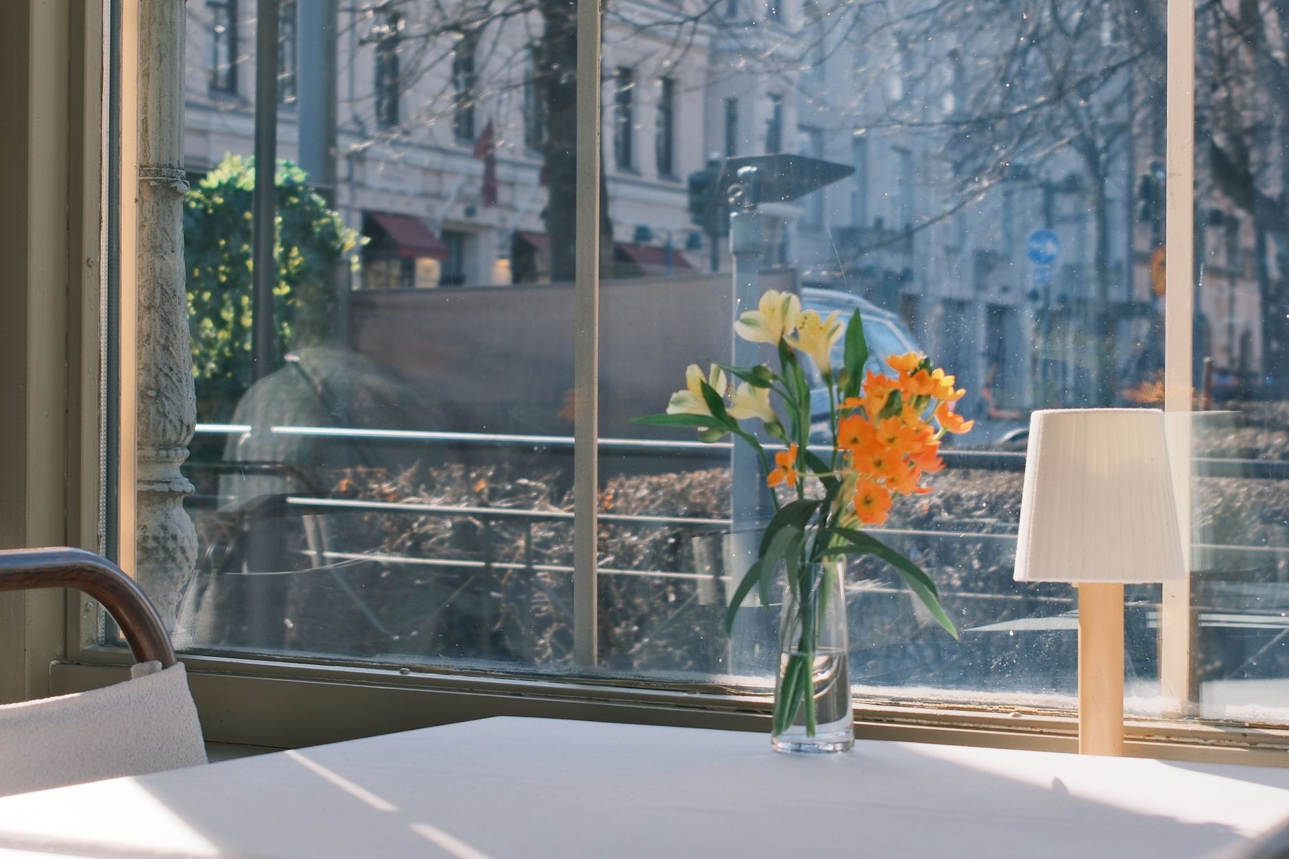 A sunlit cafe table with lilies and daffodils in a vase near a lamp and window view.