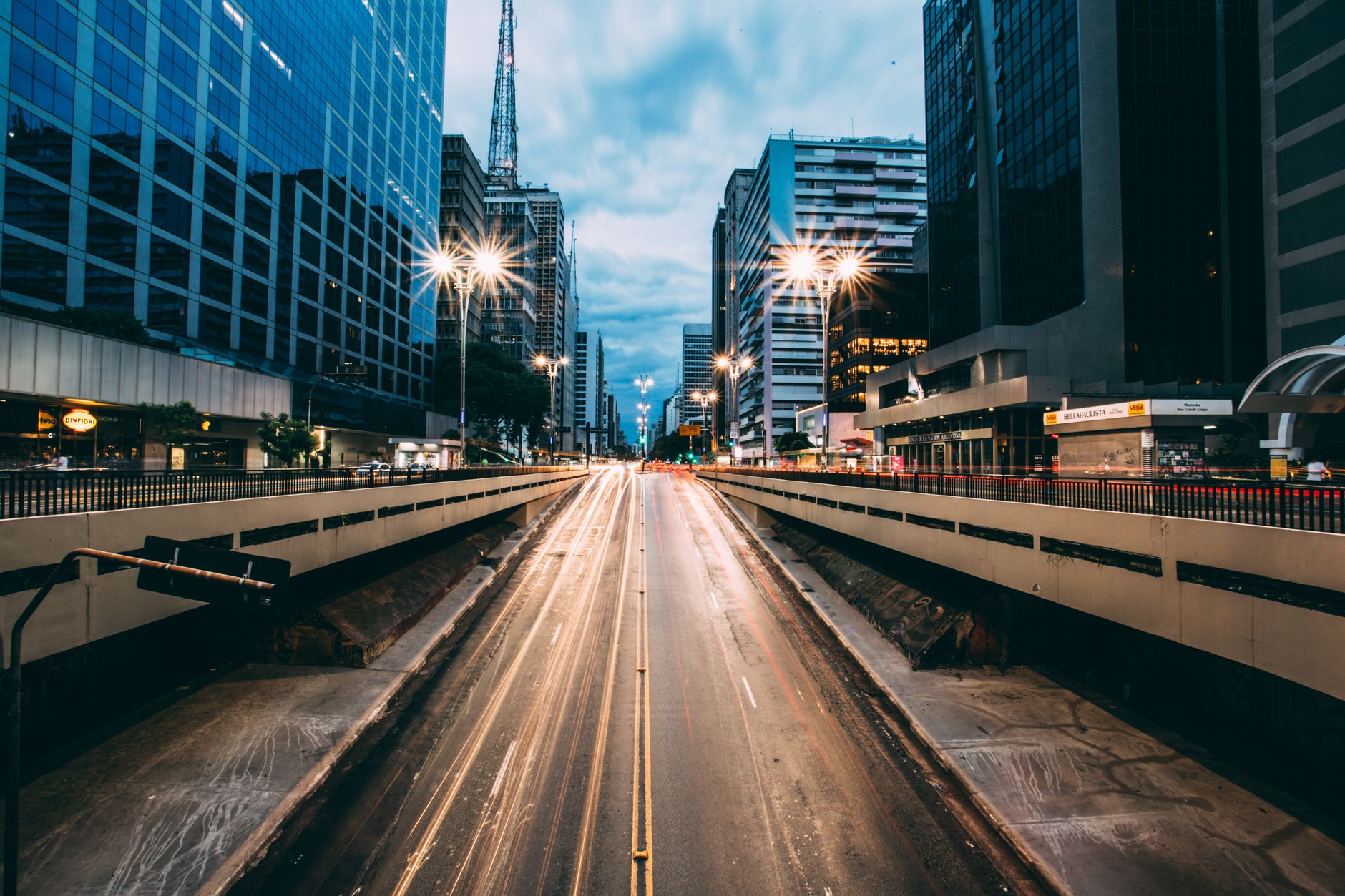 A bustling urban street scene at twilight featuring light trails from traffic, framed by tall modern buildings.