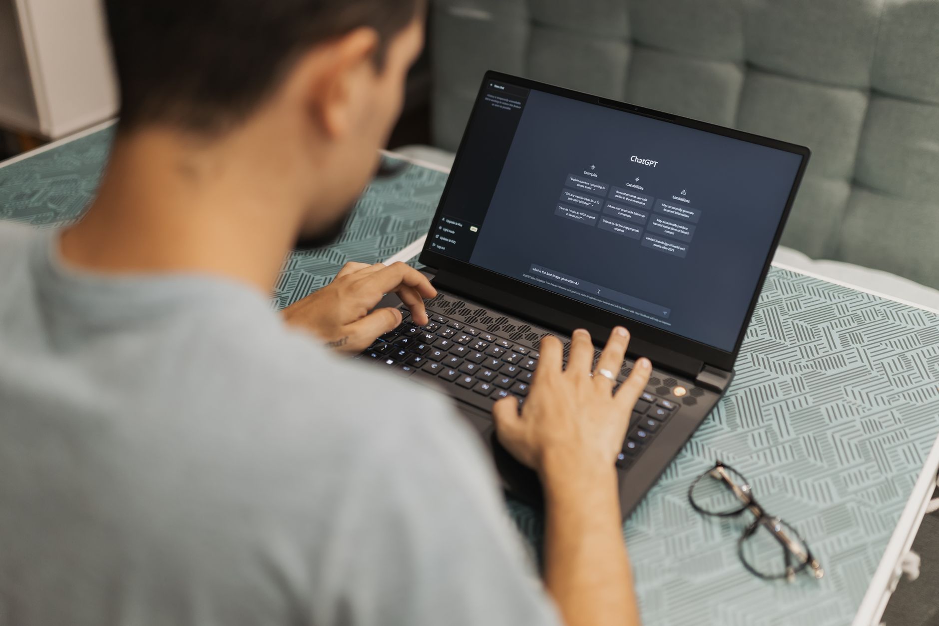Man using laptop with AI interface, typing with focused attention. Indoors with eyewear beside.