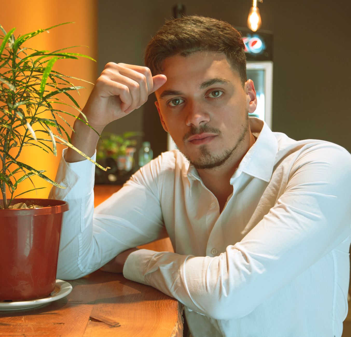 Man in white shirt with serious expression sitting by potted plant indoors.