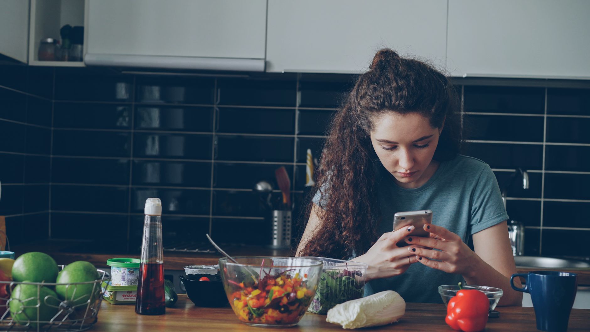A young woman checks her smartphone in a modern kitchen with fresh vegetables.