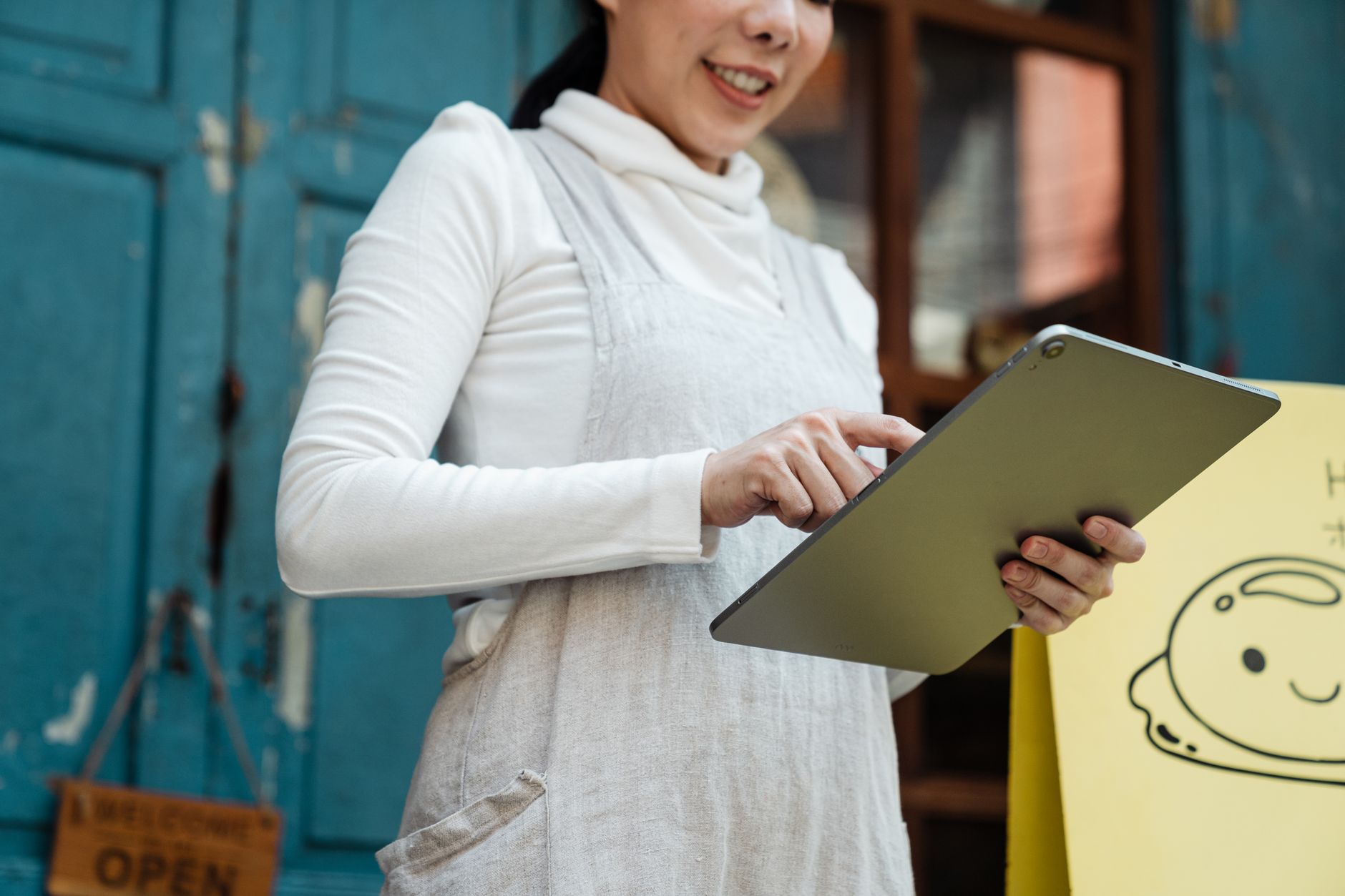 Smiling woman with a tablet in front of a shop displaying an open sign.