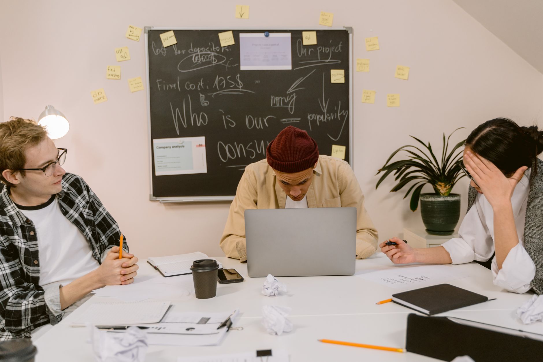 Three colleagues engage in focused discussion with laptops and notes in a modern office setting.