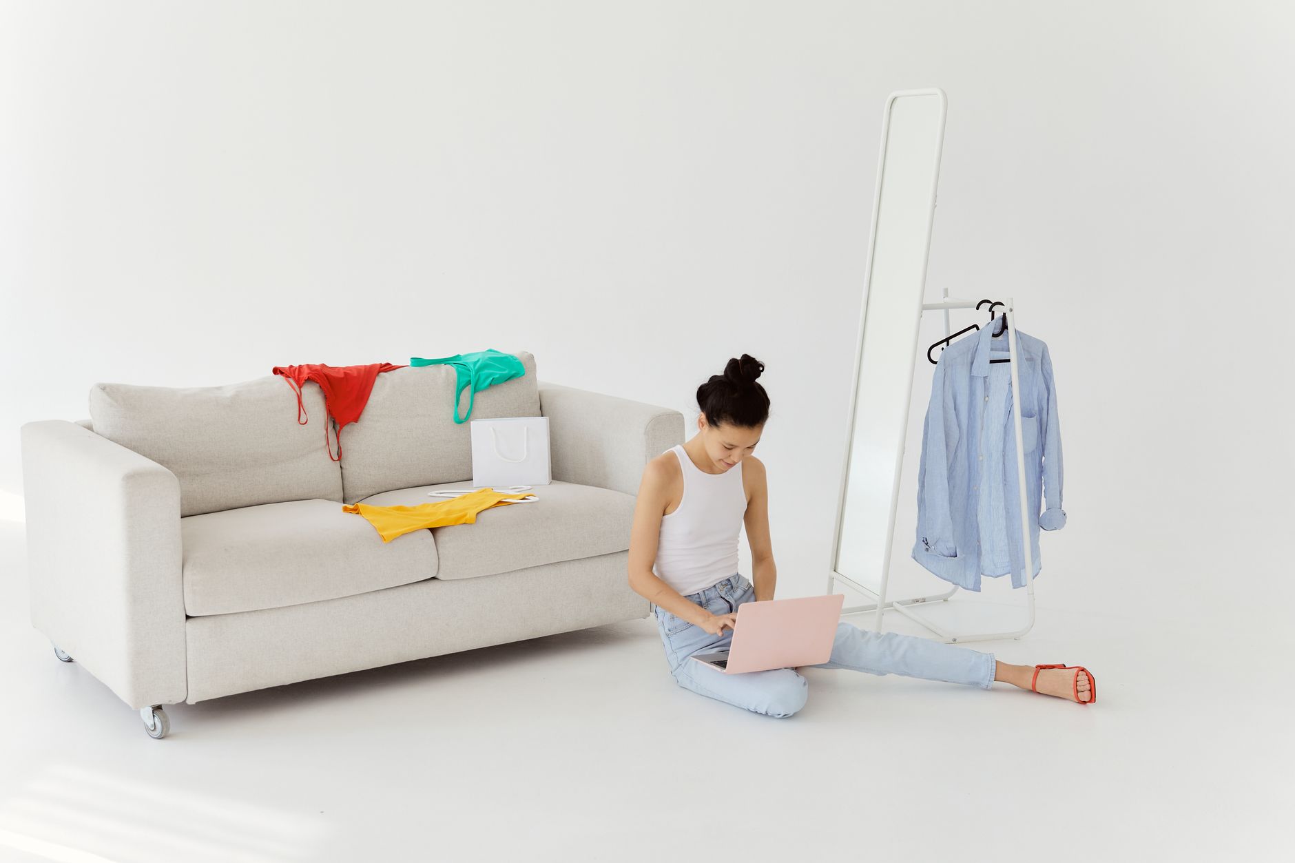 A woman in casual wear sits on the floor using a laptop next to a sofa and clothes rack.