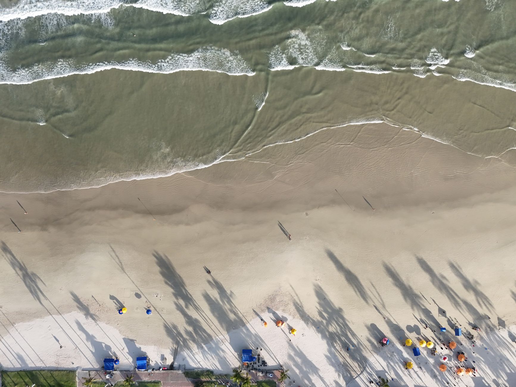 Serene drone view of a sandy beach with palm shadows and gentle waves lapping the shore.