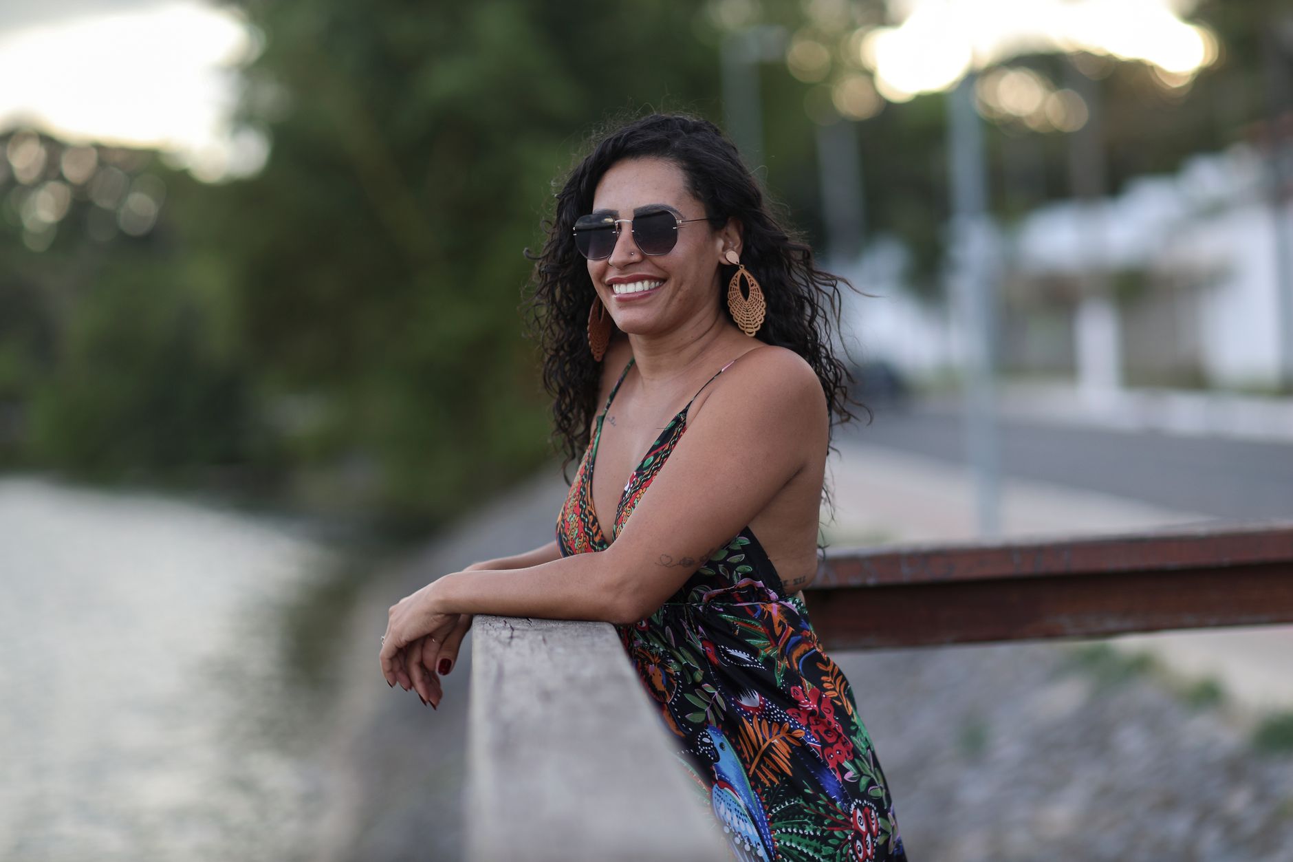 Woman in sunglasses leaning on railing by riverside, enjoying outdoor scenery.
