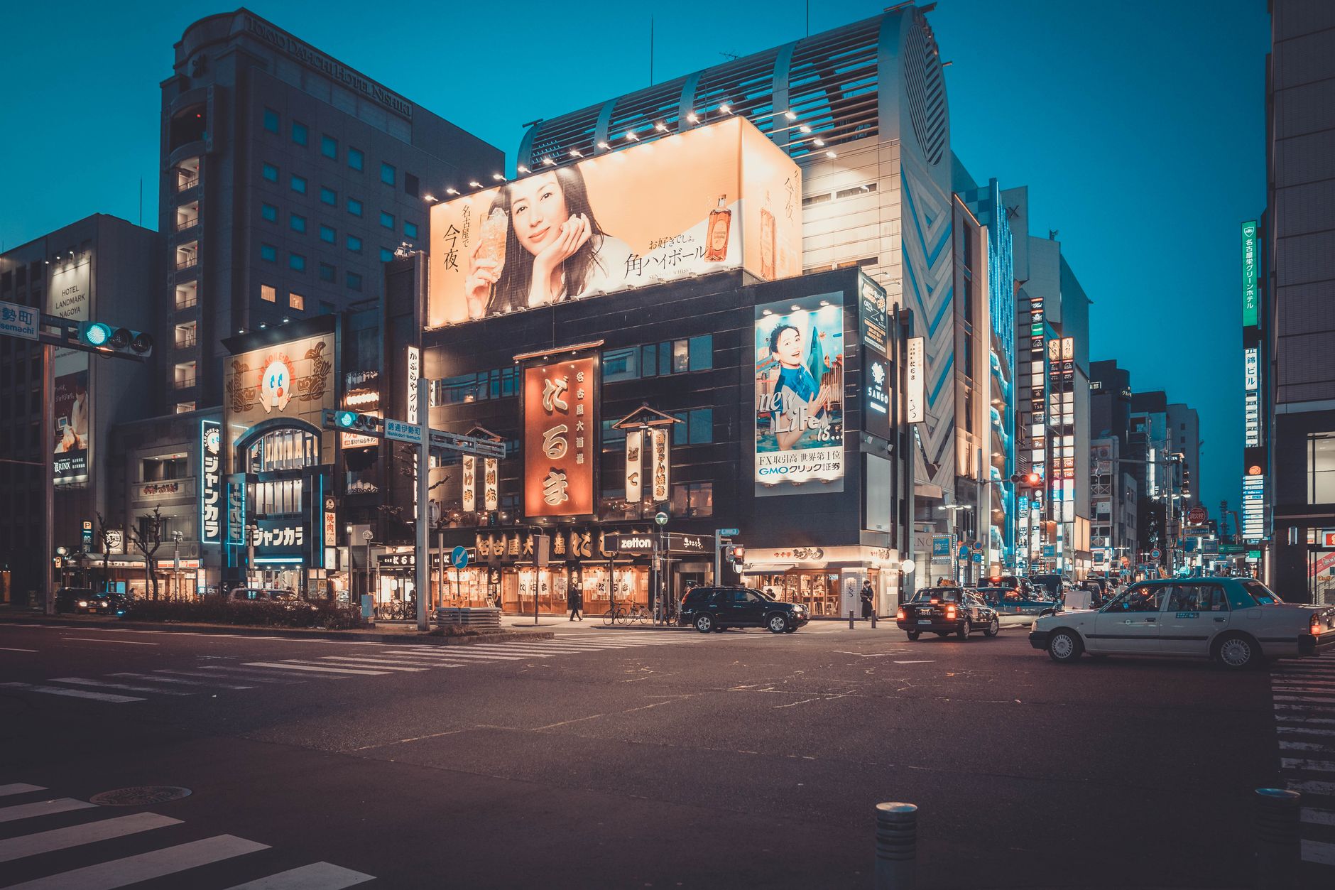 Bustling urban intersection with colorful neon signs and advertisements at twilight, showcasing modern city life.
