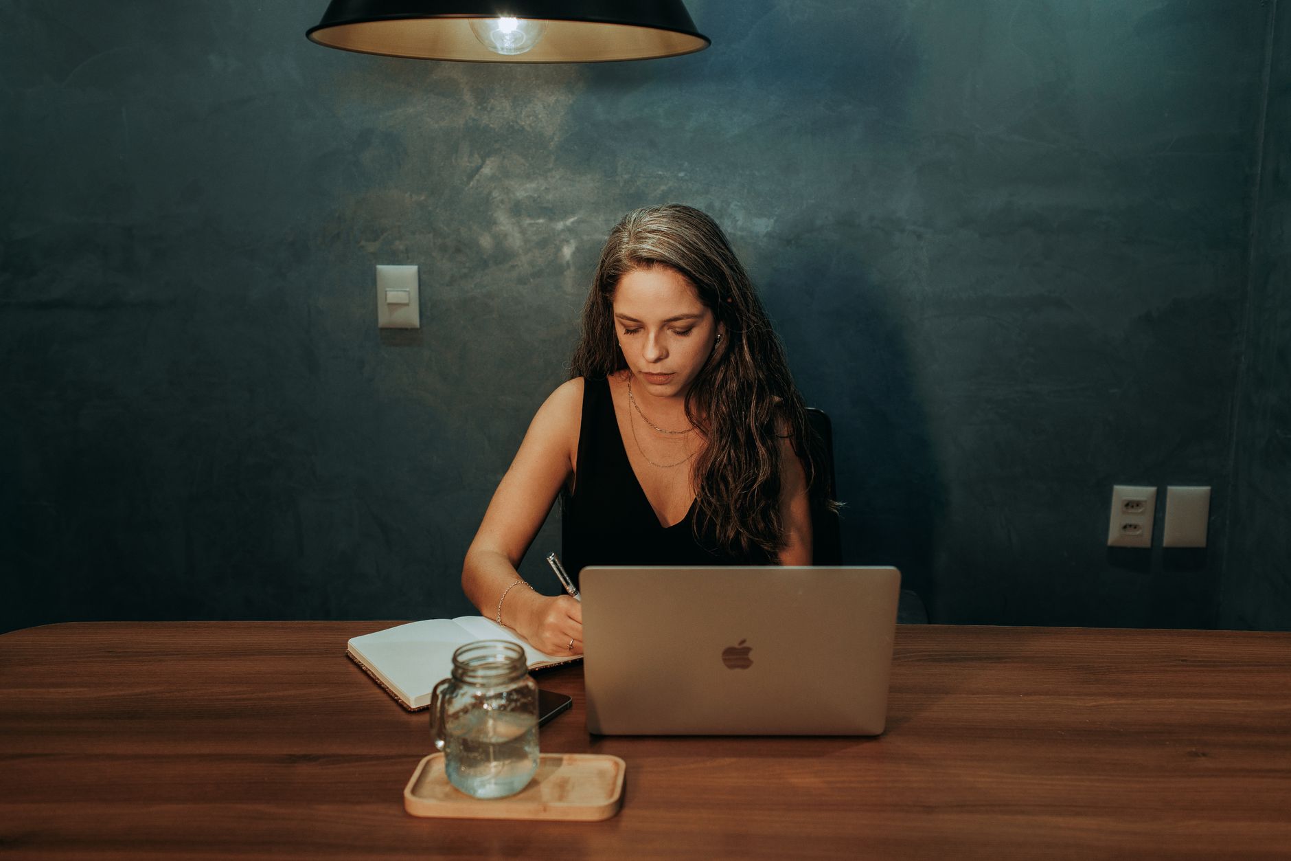 Woman engaged in work at a home office with a laptop and notebook under warm lighting.