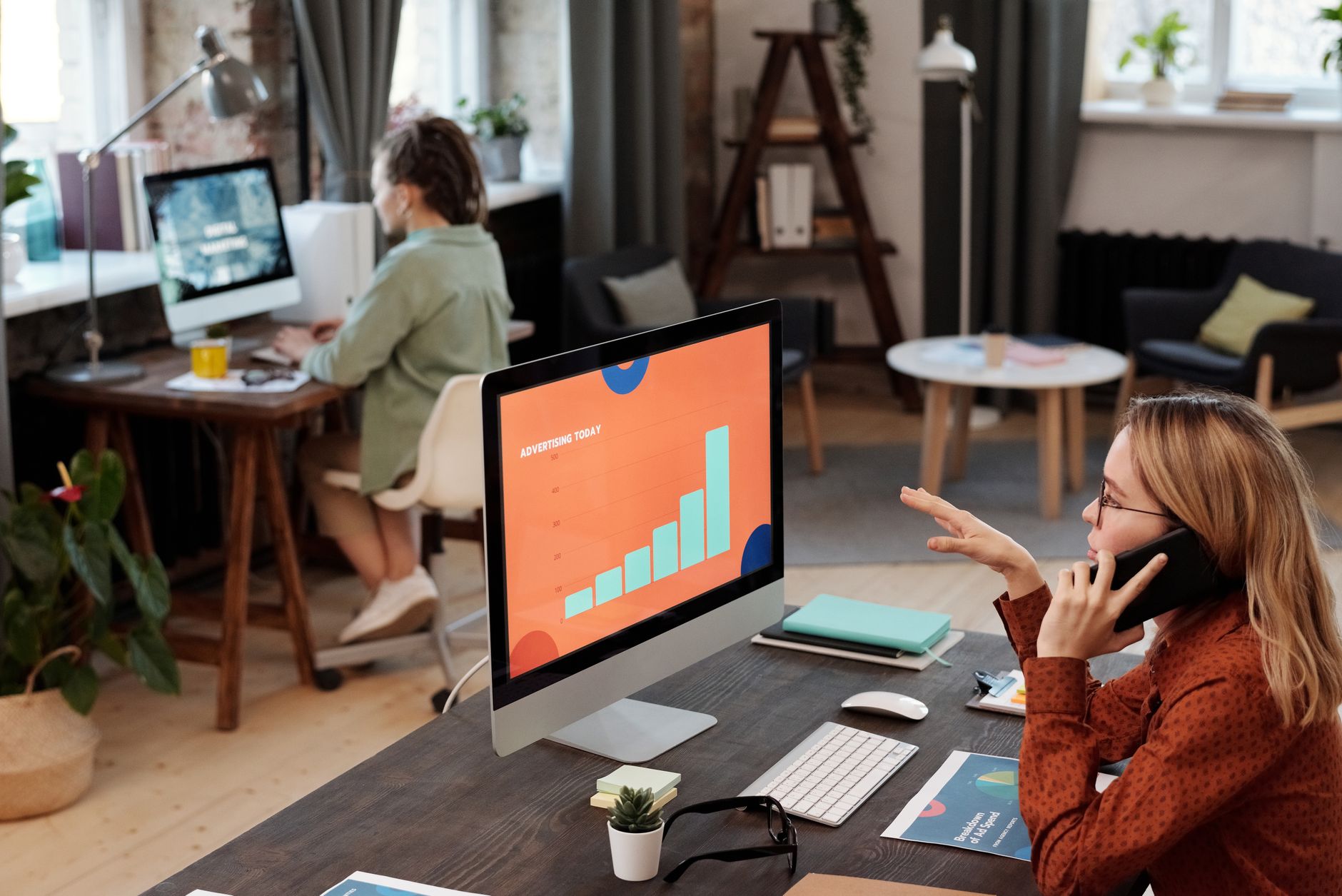 Two business professionals engaged in work with computers and phone in a modern office setting.