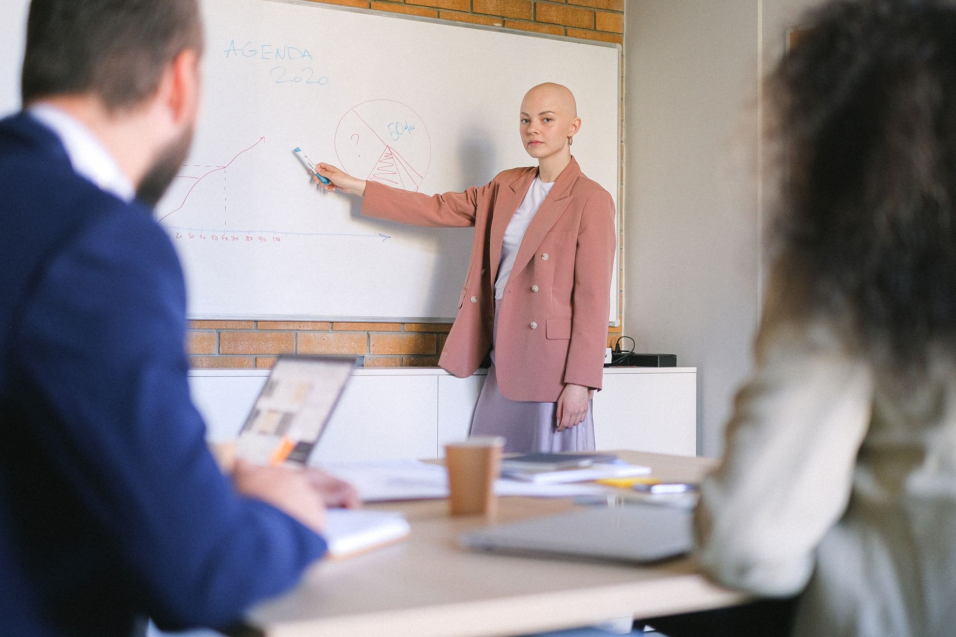 Serious woman pointing with marker at whiteboard with diagrams and charts while explaining business plan to colleagues in conference room