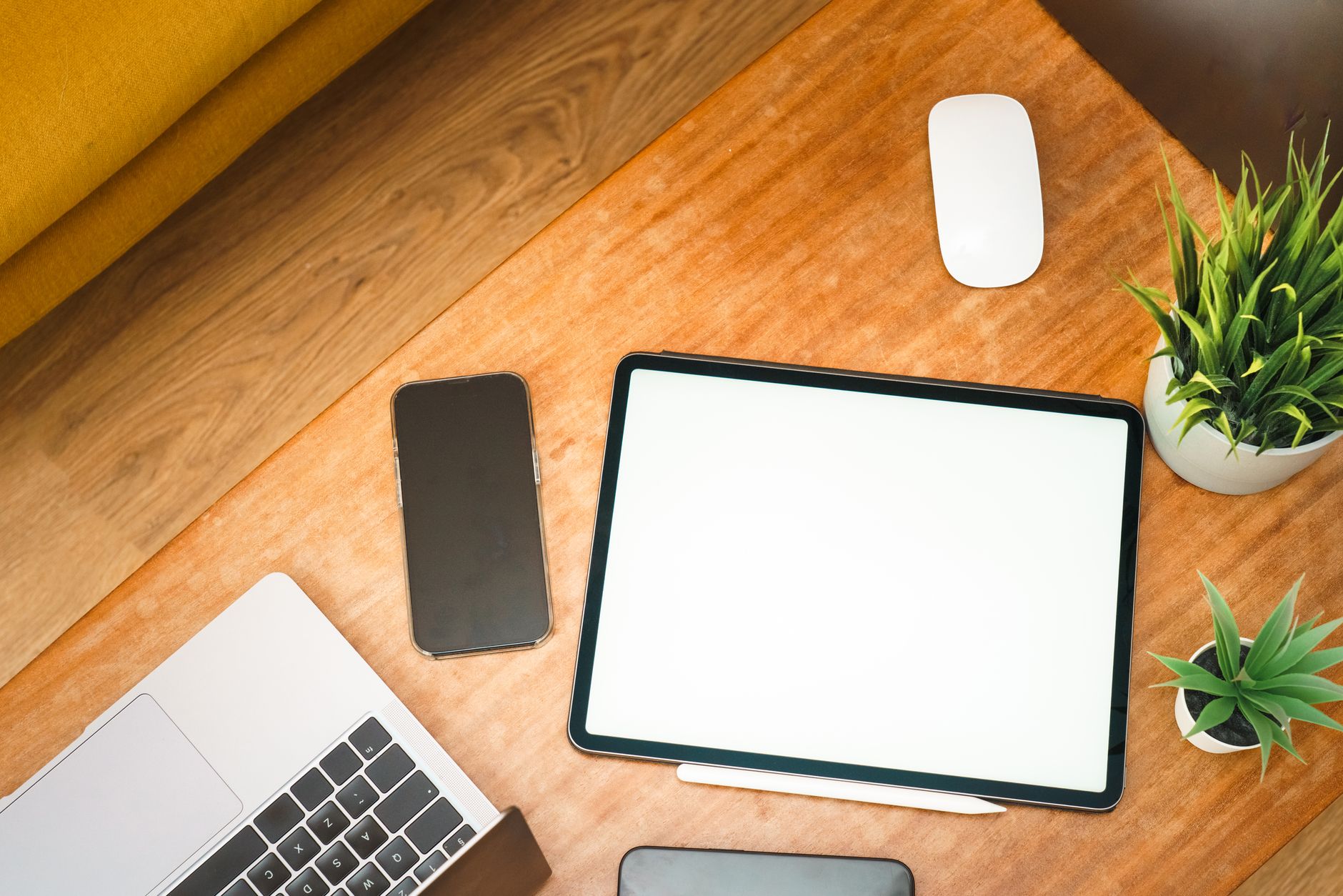 Top view of a stylish workspace featuring a laptop, tablet, smartphone, and plants on a wooden desk.