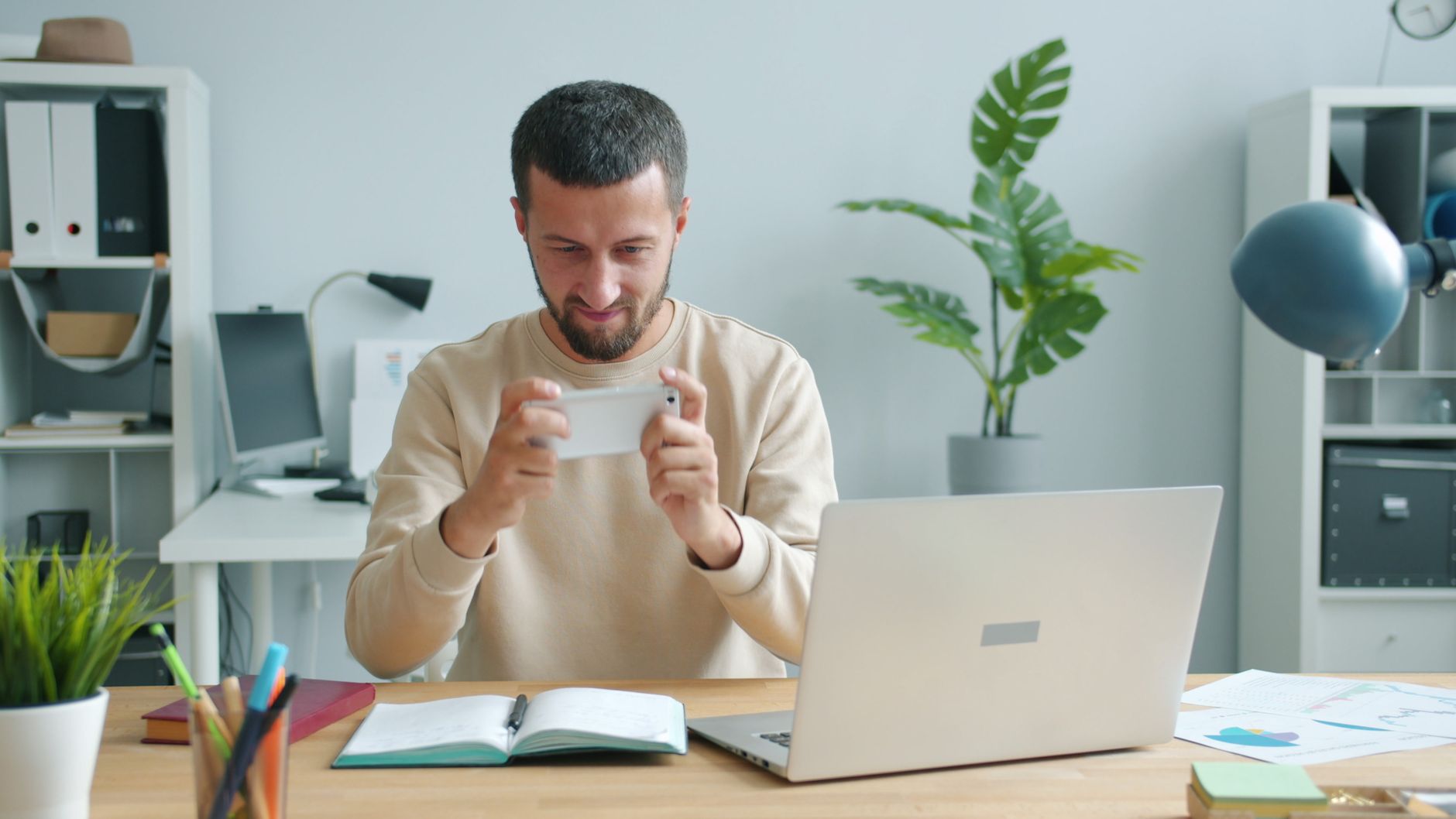 Focused man using smartphone at a desk with laptop in a modern office setting.