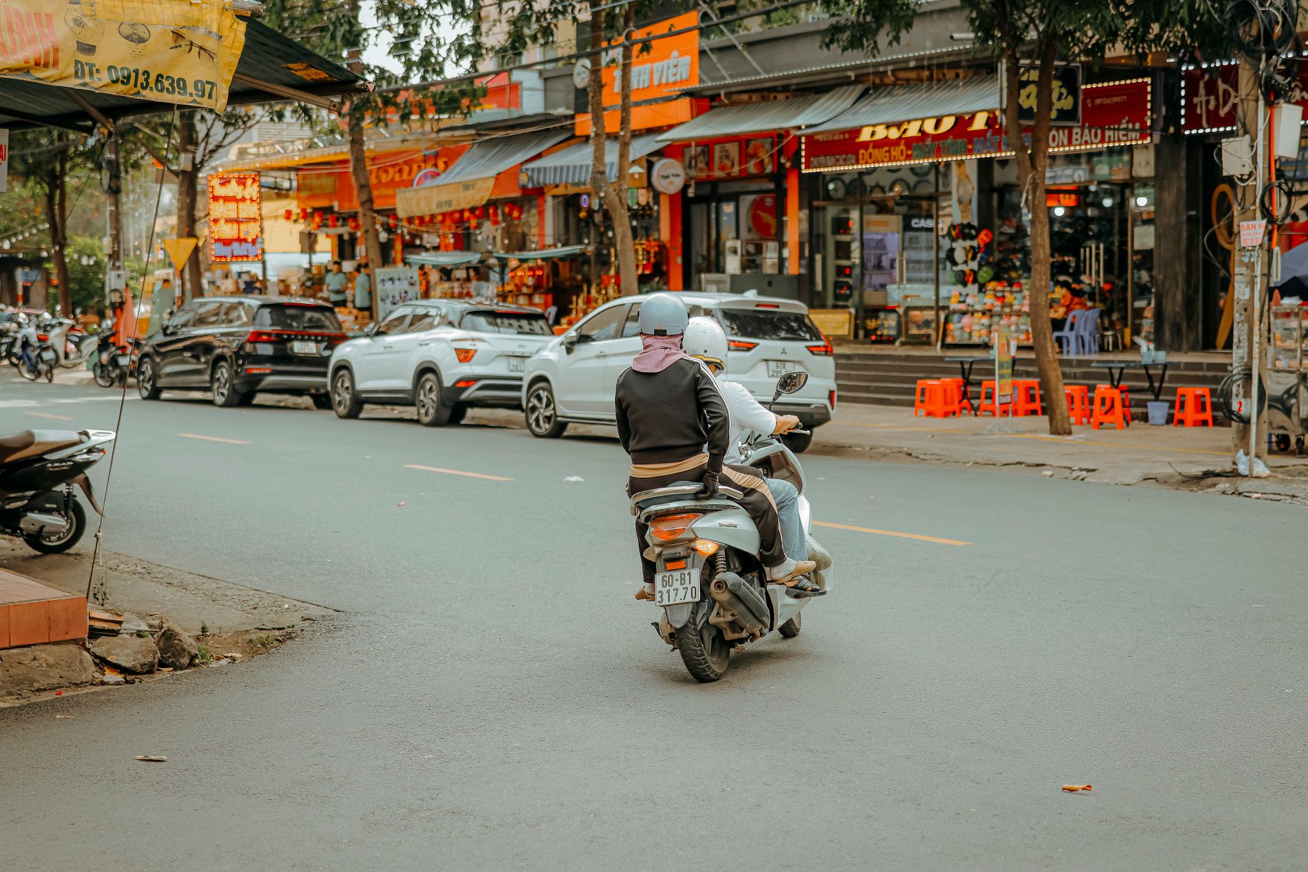 Two riders on a motorbike navigate a bustling city street lined with shops.