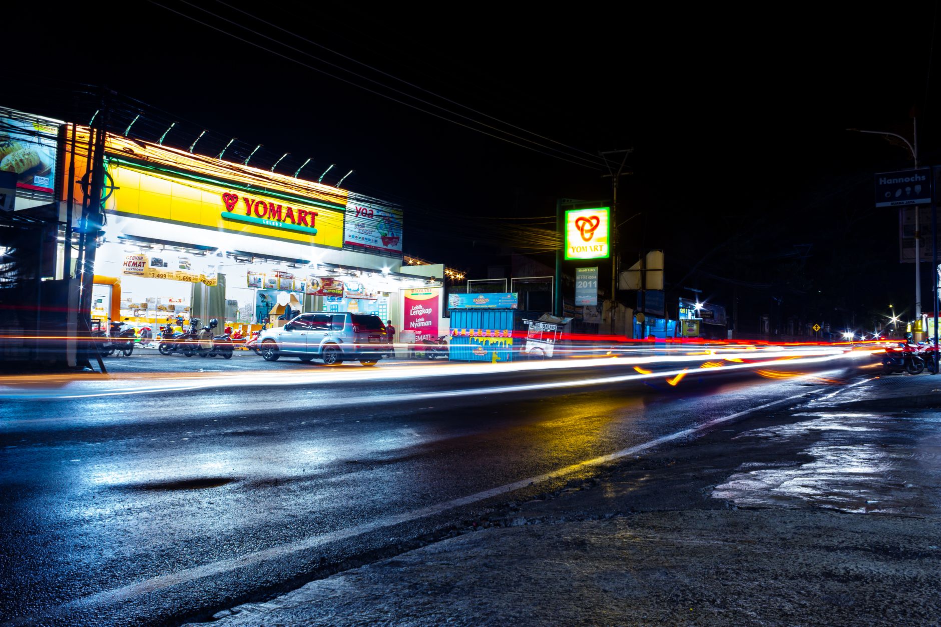 A lively city street scene in West Java showcasing long exposure light trails and a shopping mart at night.
