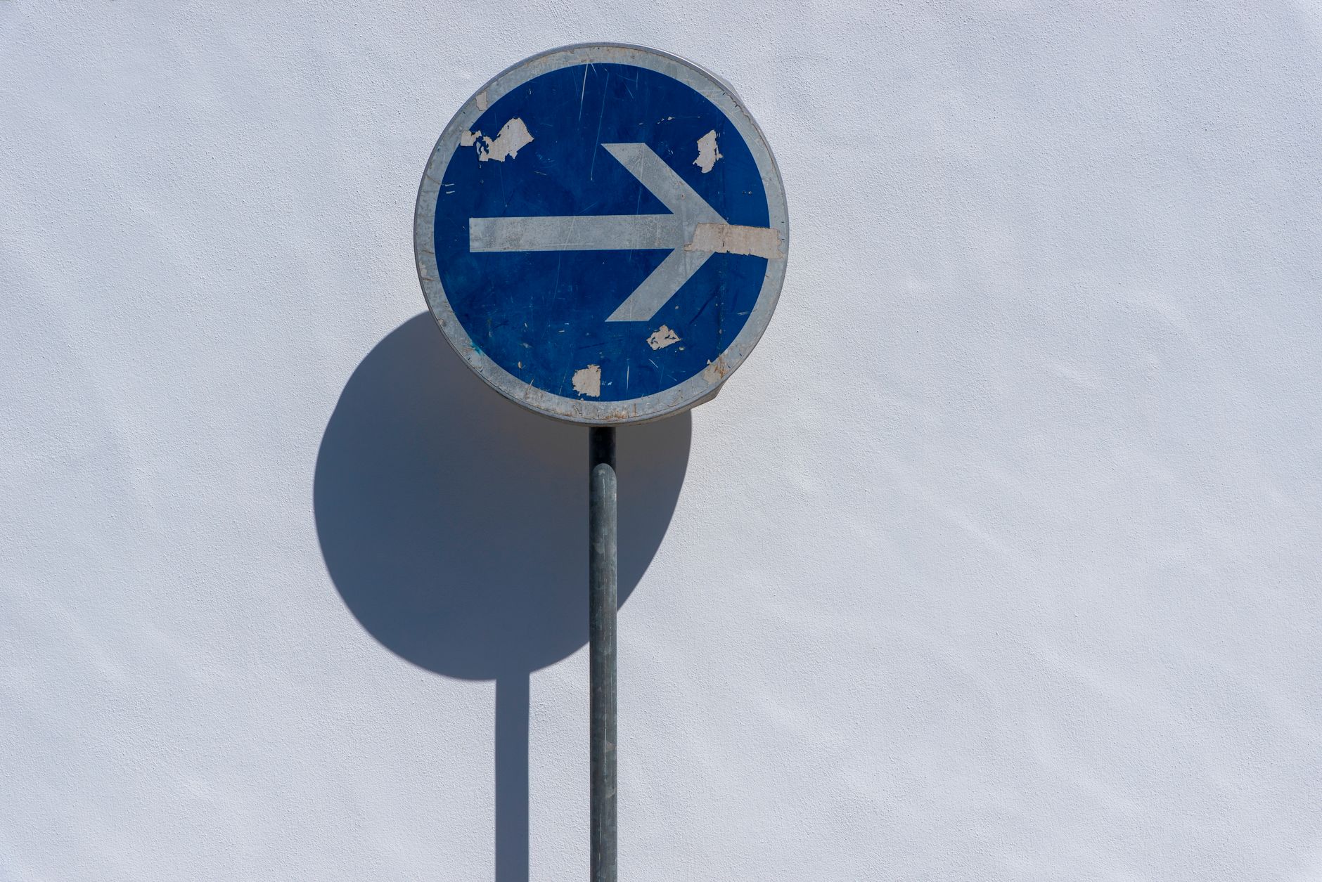 Weathered blue road sign with right arrow on a sunny day against a white wall.