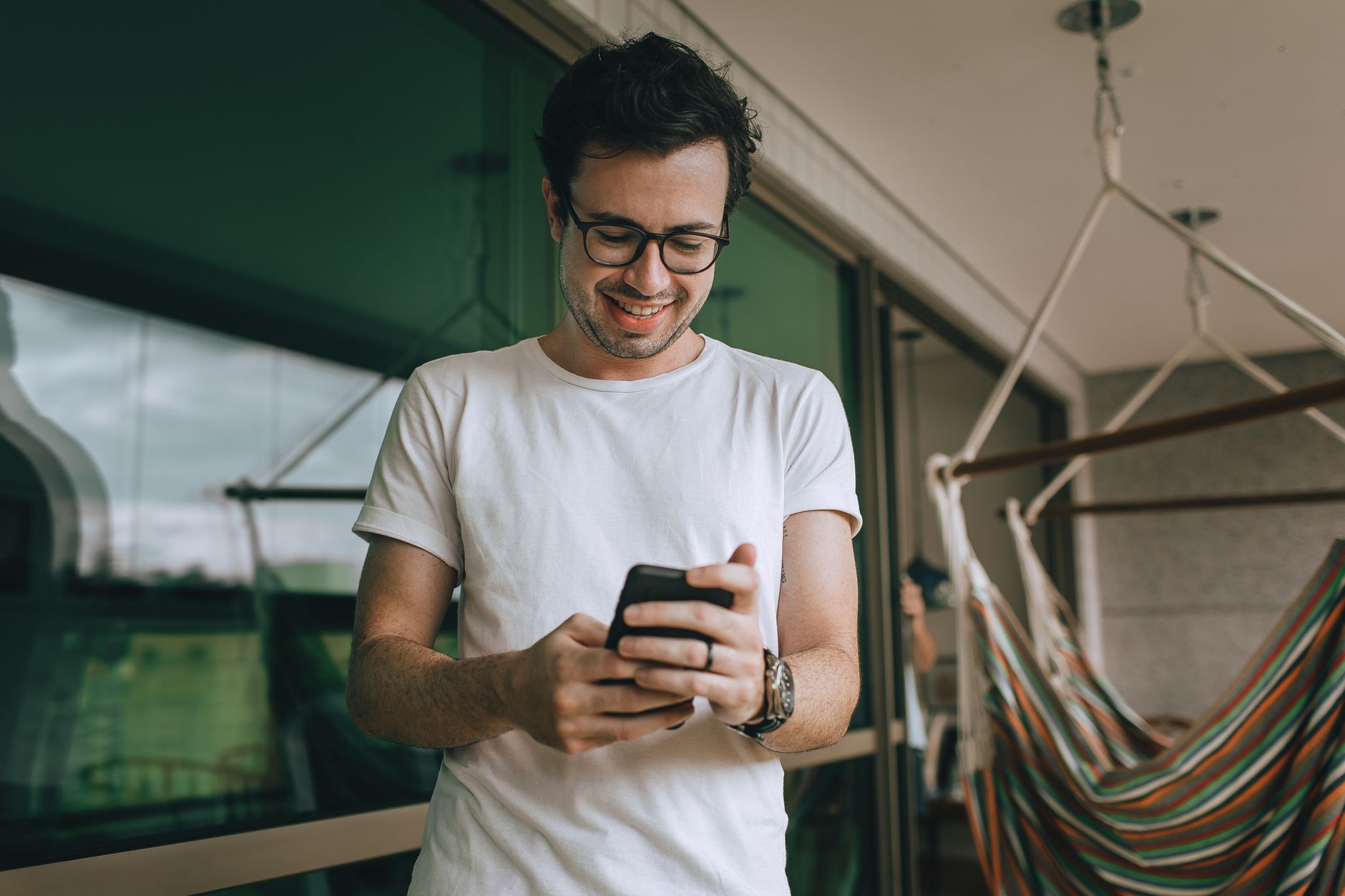 Casual man in glasses smiling while messaging on a smartphone outdoors.