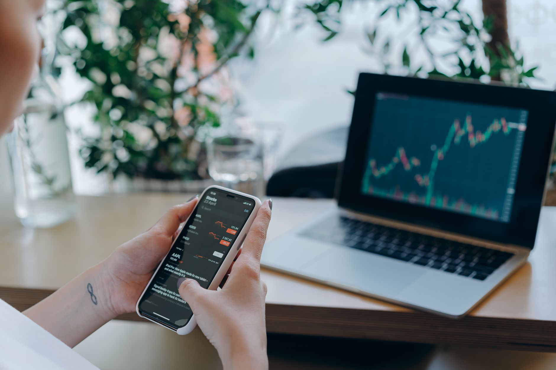 Businesswoman reviewing financial data on smartphone alongside laptop displaying graphs in modern workspace.