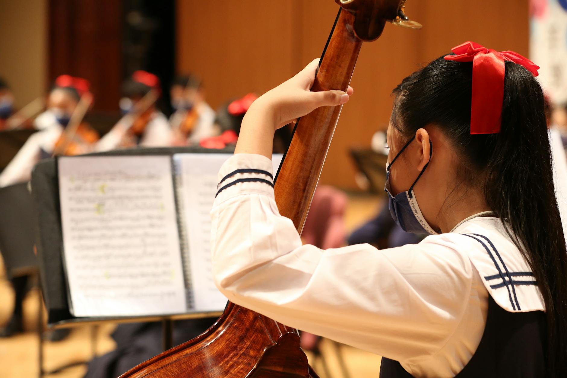 A young girl performs cello in an indoor school orchestra setting.