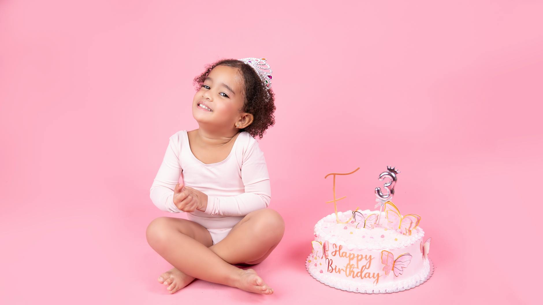 Smiling child in tiara with birthday cake and pink backdrop.
