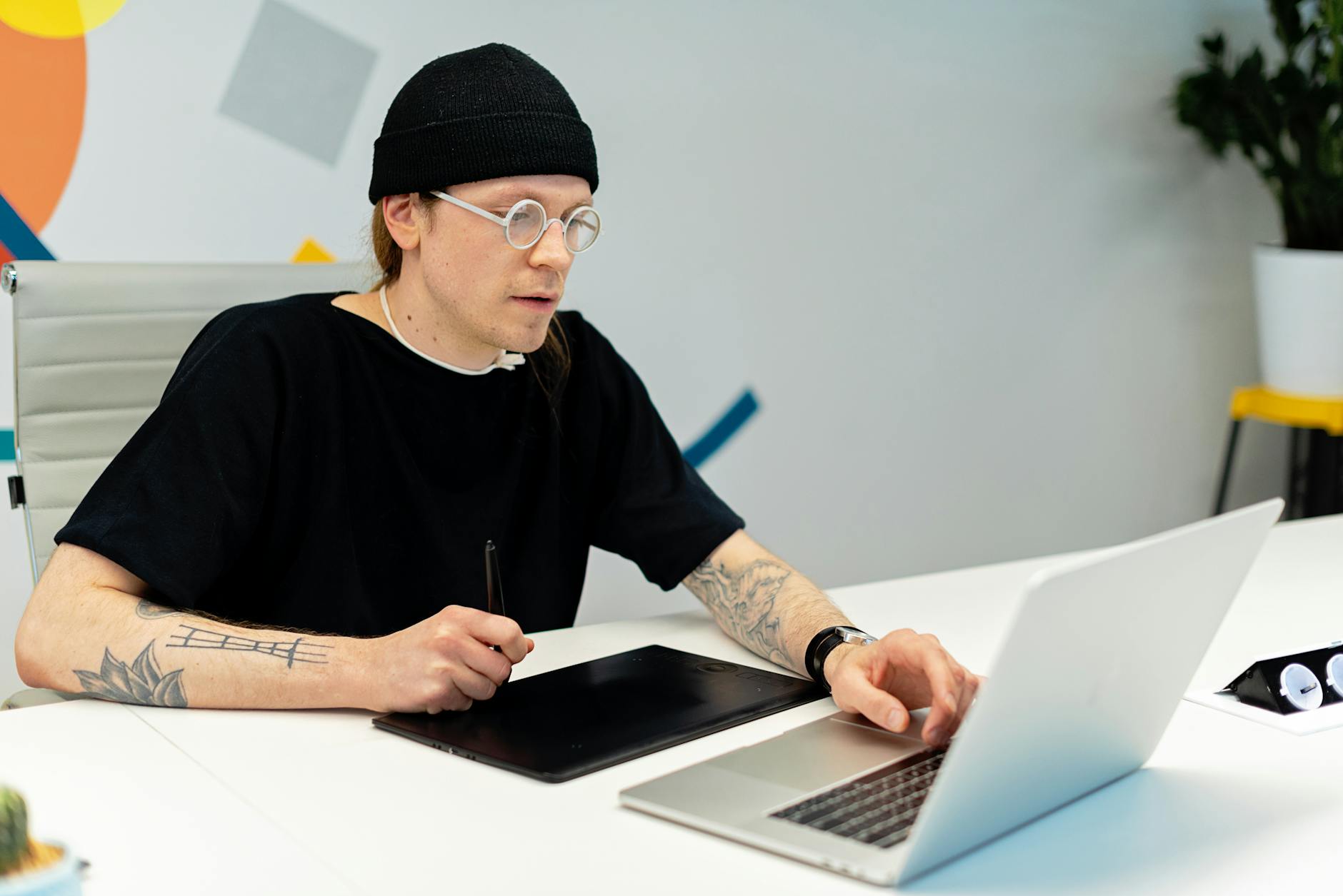 Tattooed designer working on a laptop and tablet in a stylish office setting.