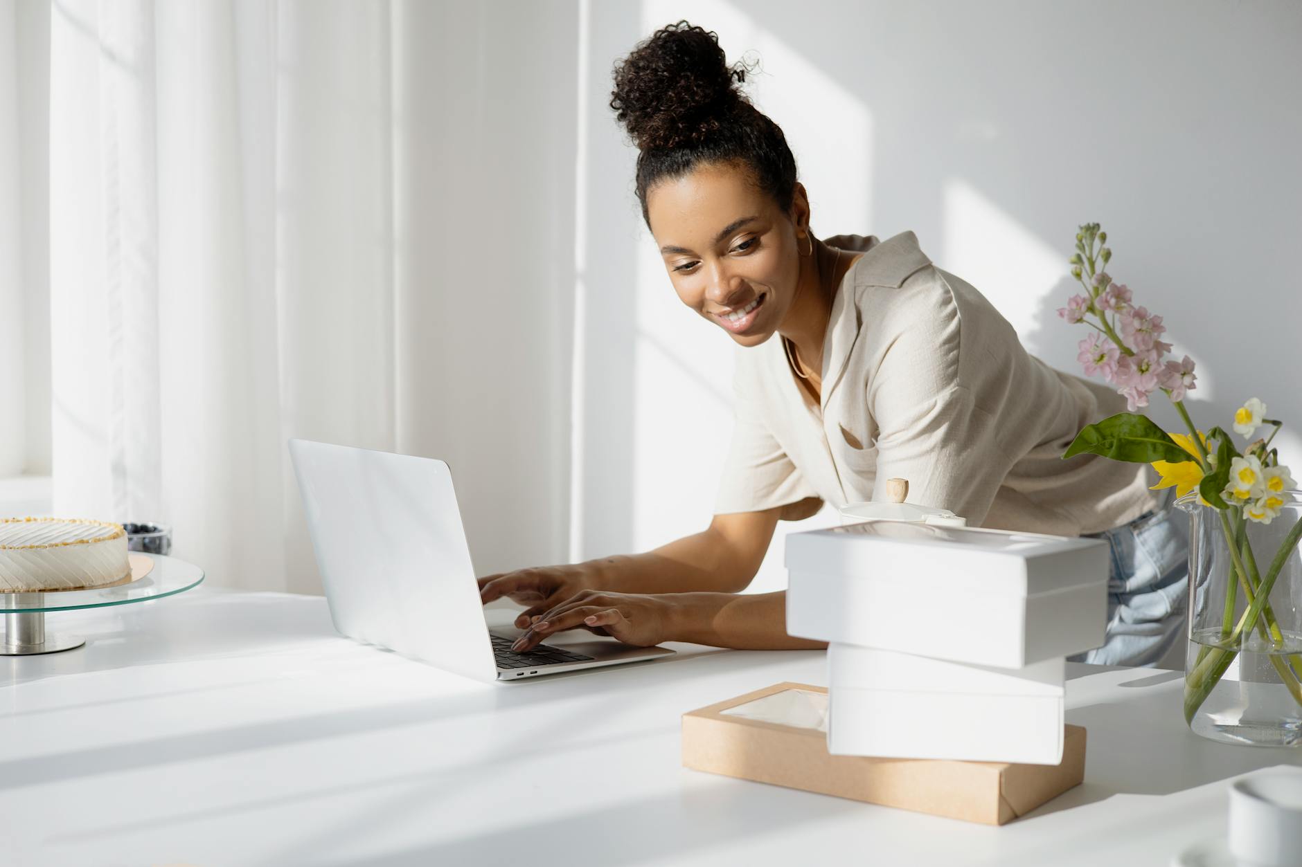African American woman working on a laptop with boxes and flowers in a bright room.