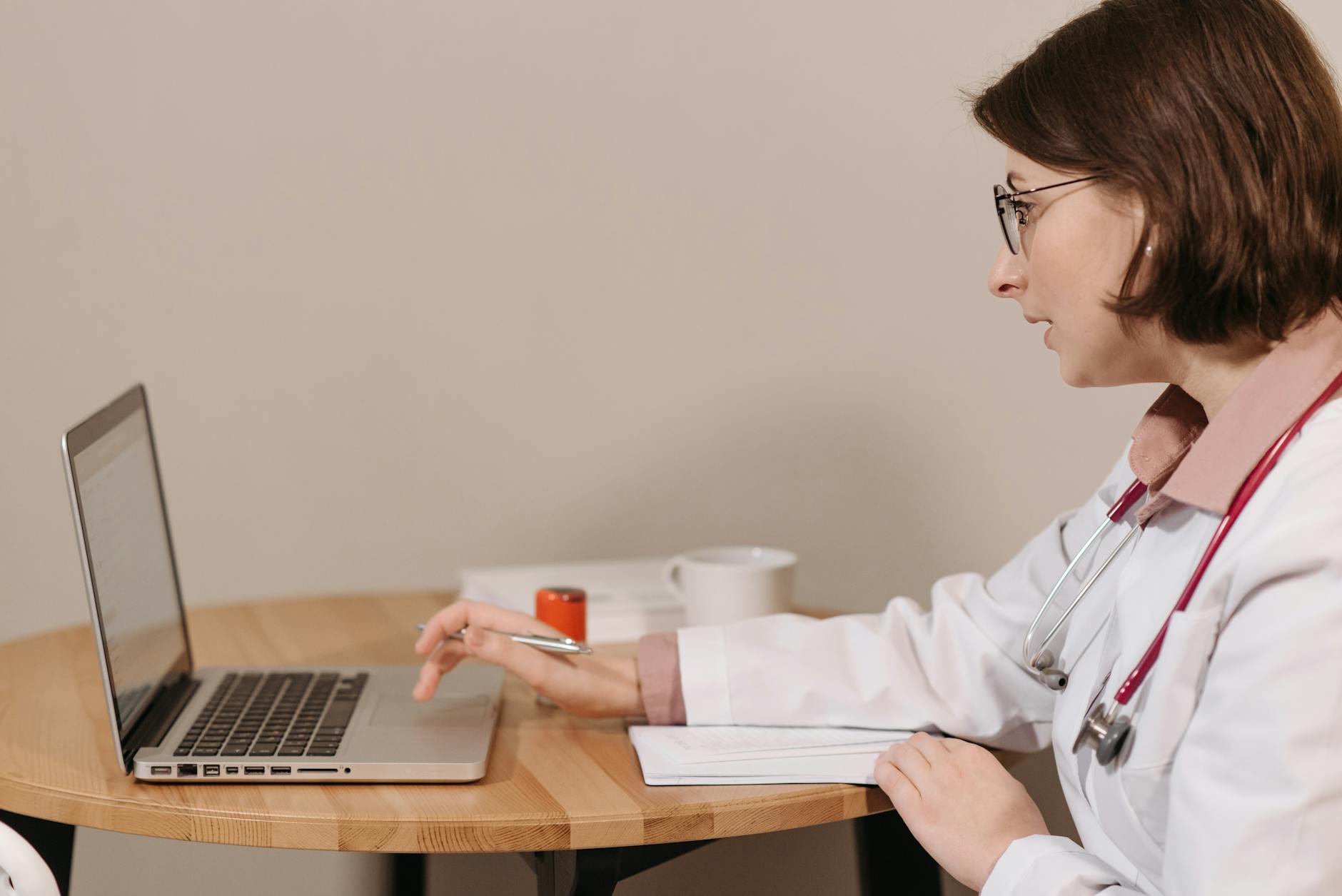 A female doctor in a white coat using a laptop while writing notes at a wooden table.