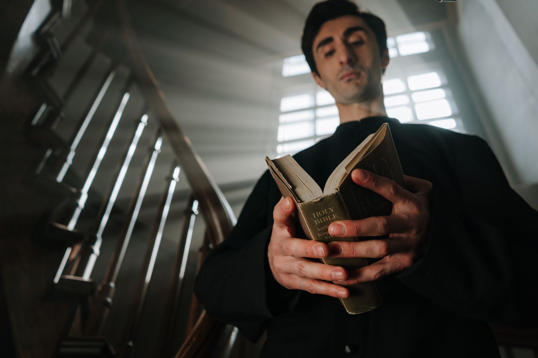 A priest in black robes reads a Bible by a sunlit staircase indoors.