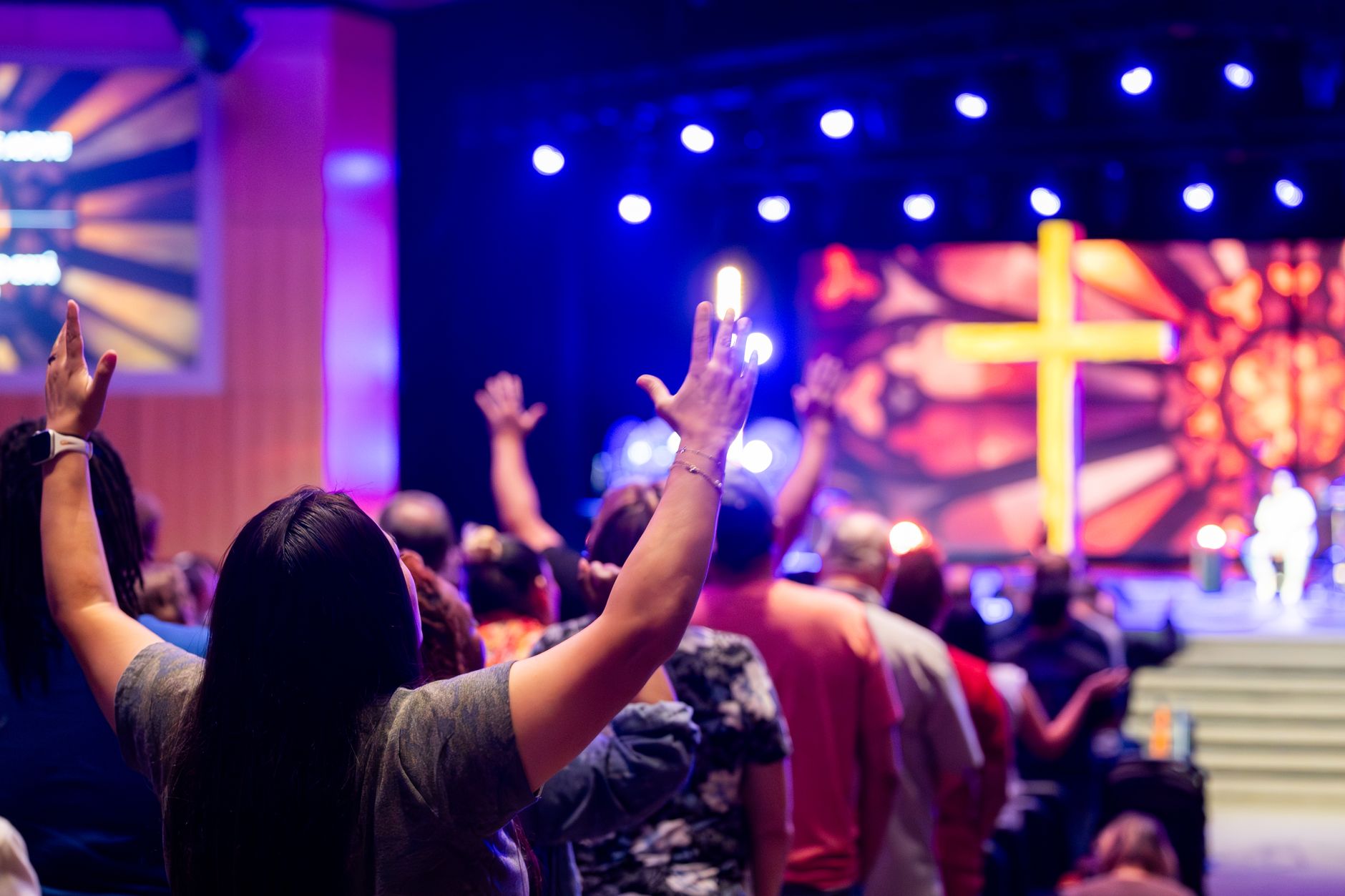 People engaged in a vibrant worship service, raising hands in faith under colorful stage lights and a prominent cross.