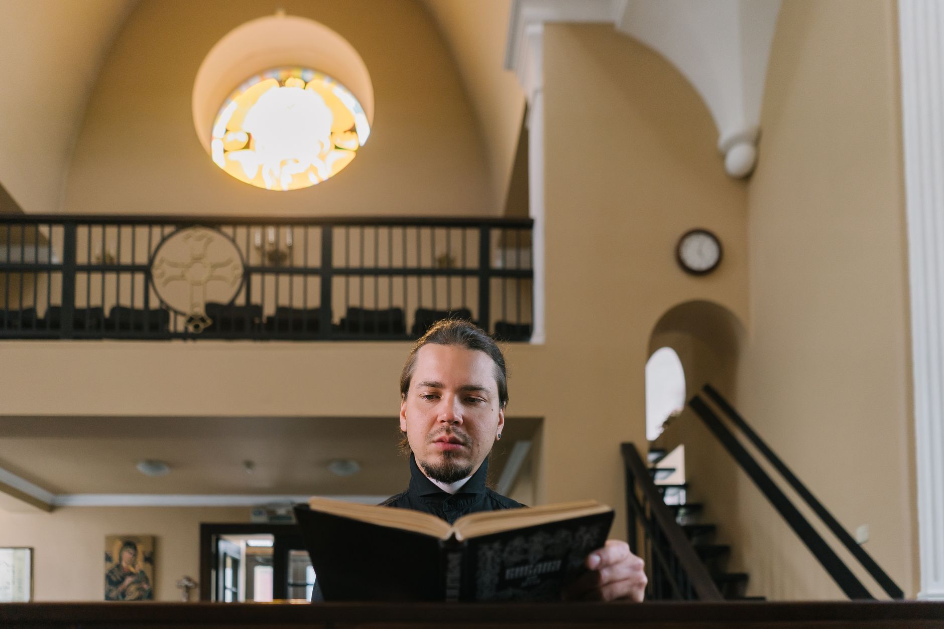 A priest reads a holy book inside a church under a stained-glass window.