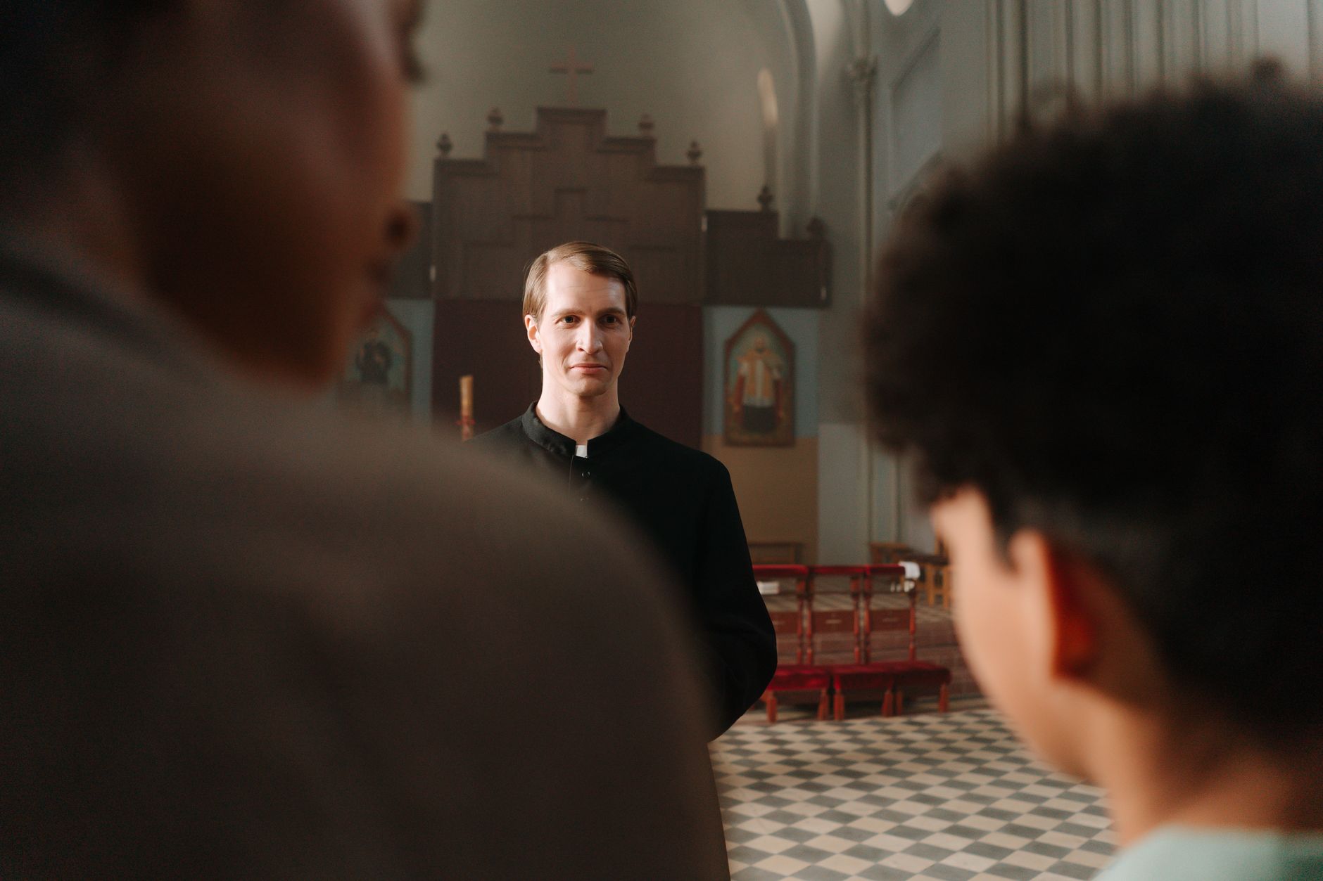 A priest in a cassock listening to churchgoers inside a serene church setting.