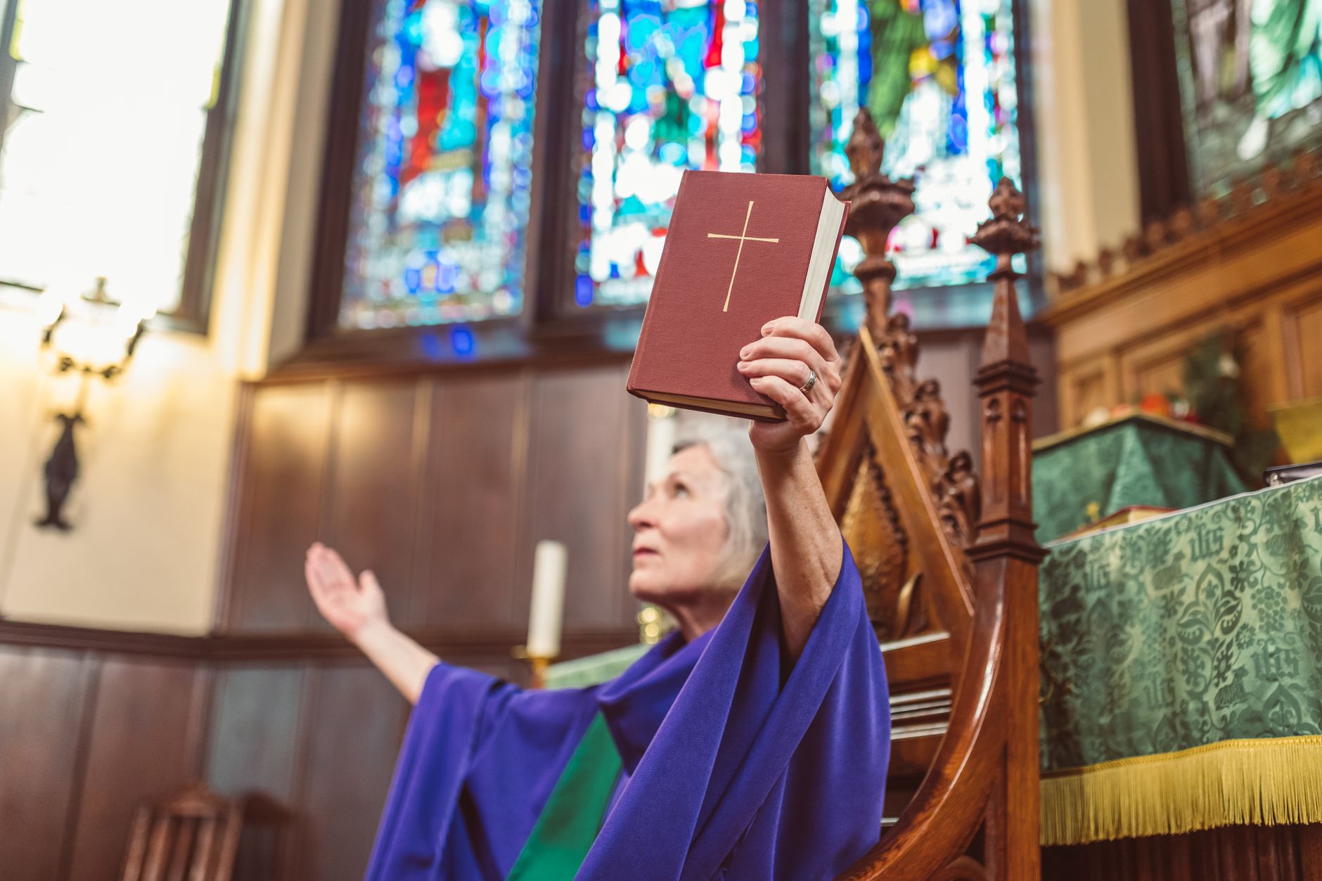An elderly priest praying inside a cathedral, holding a Holy Bible near stained glass windows.
