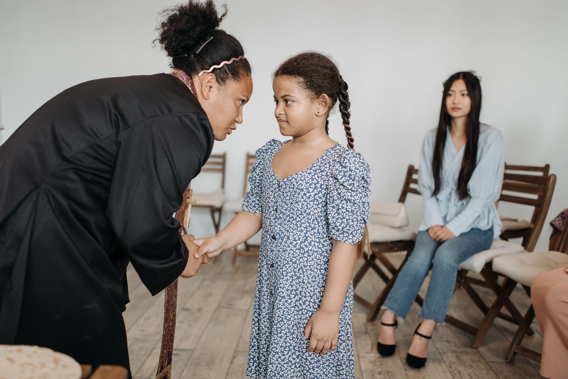 A clergy member interacts warmly with a young girl while others look on in a meeting room setting.