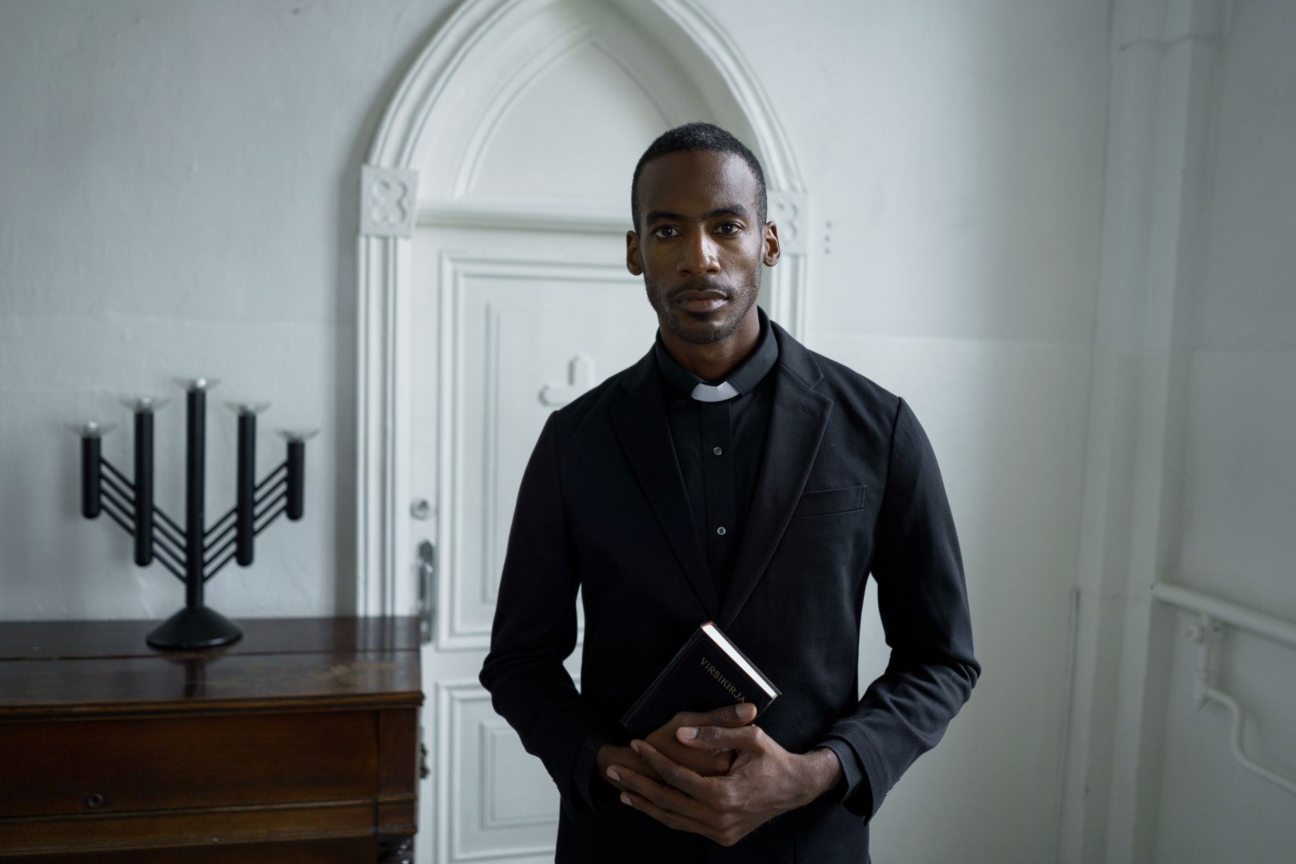 A solemn priest in black attire holds a Bible, standing inside a church.