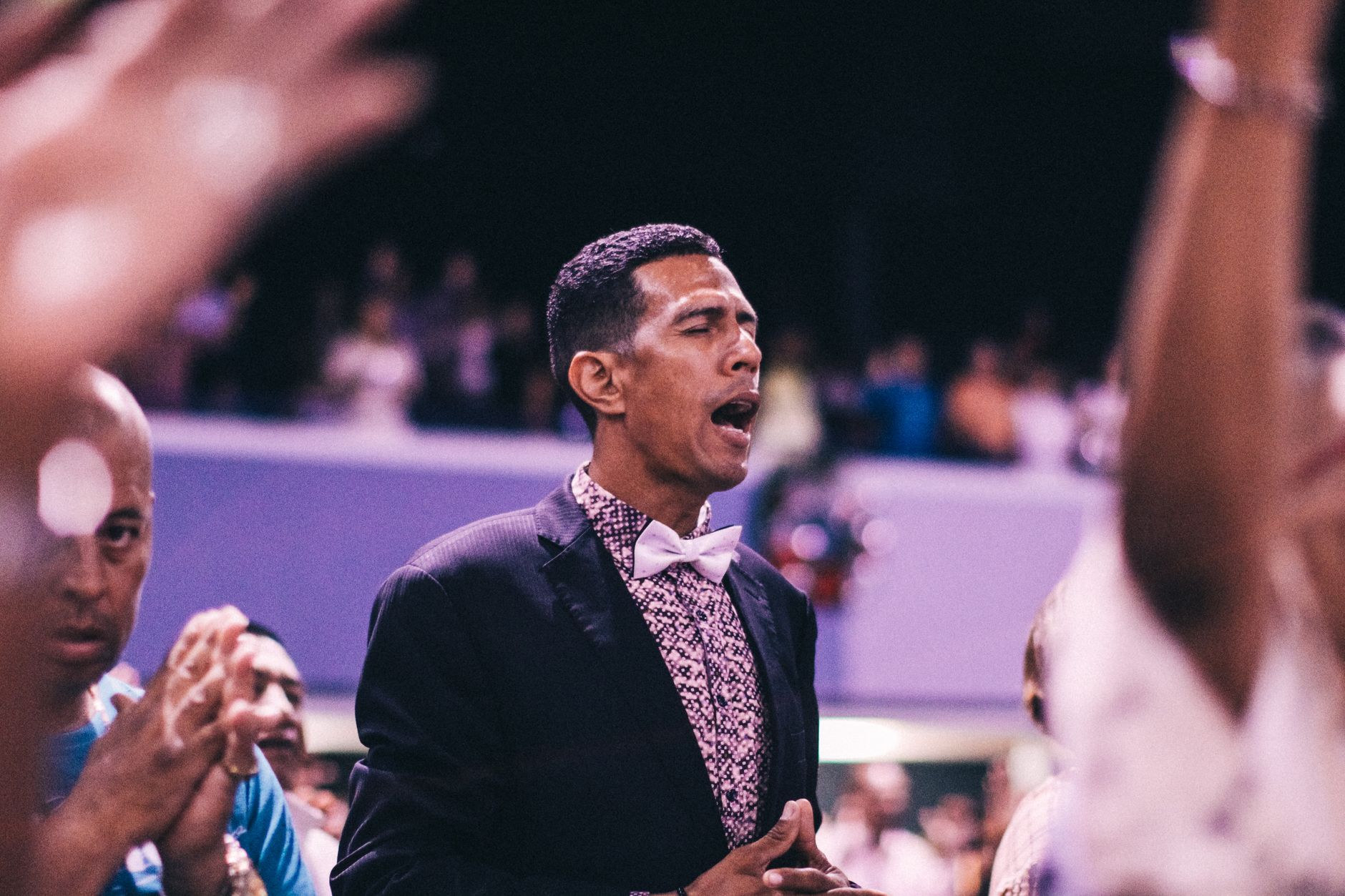 Stylish man wearing bow tie passionately praying at an indoor event with a crowd.