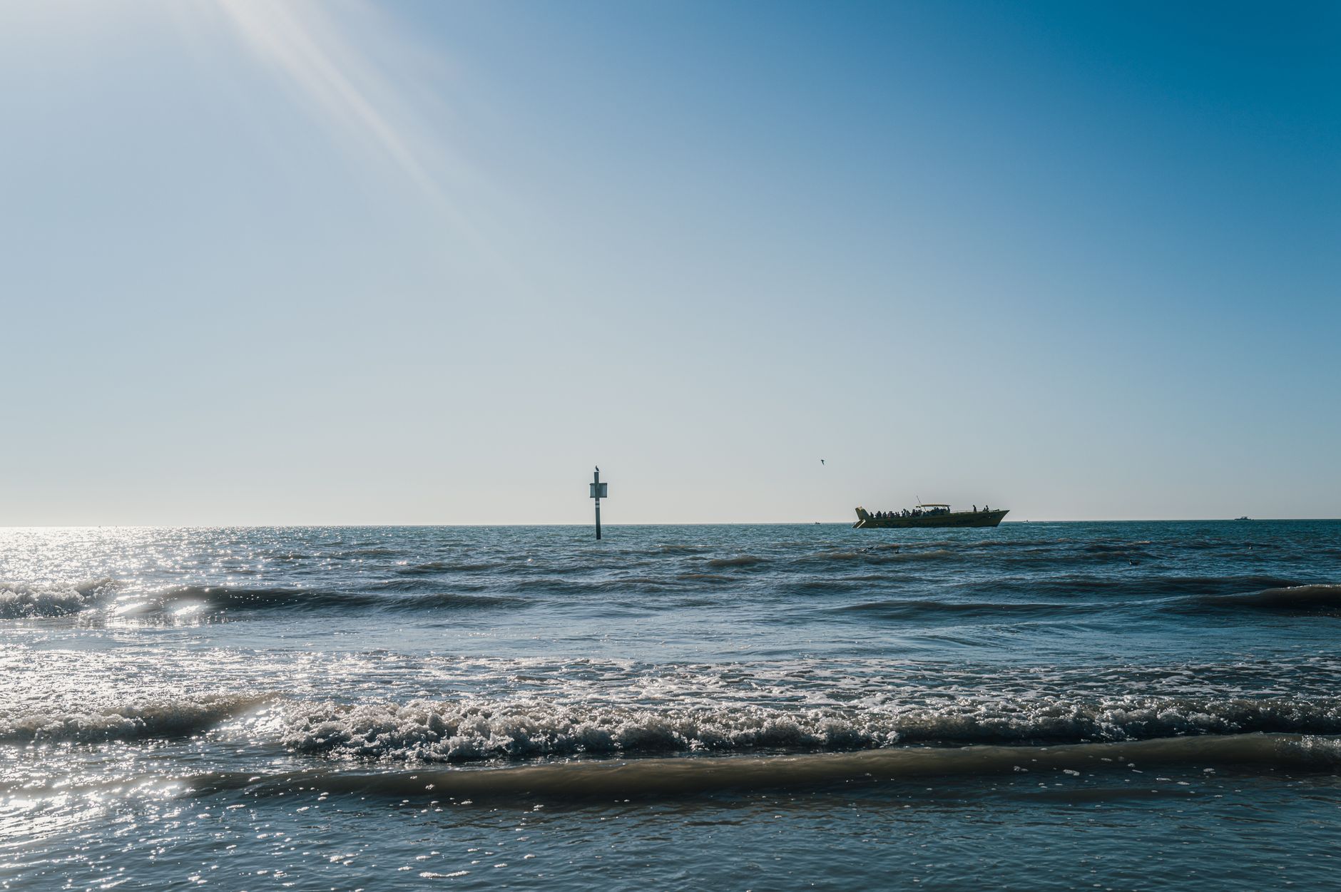 Calm ocean landscape with sparkling water, a distant boat, and navigation beacon under a clear sky.