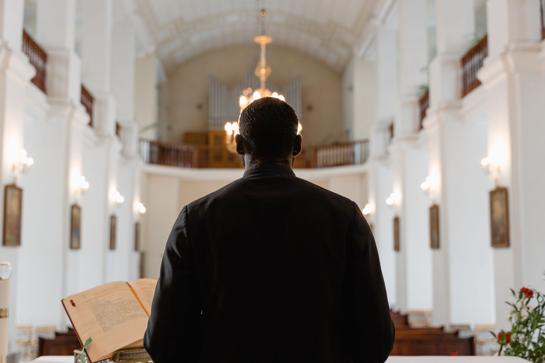 A priest stands before a congregation in a grand church, delivering a sermon with an open Bible.