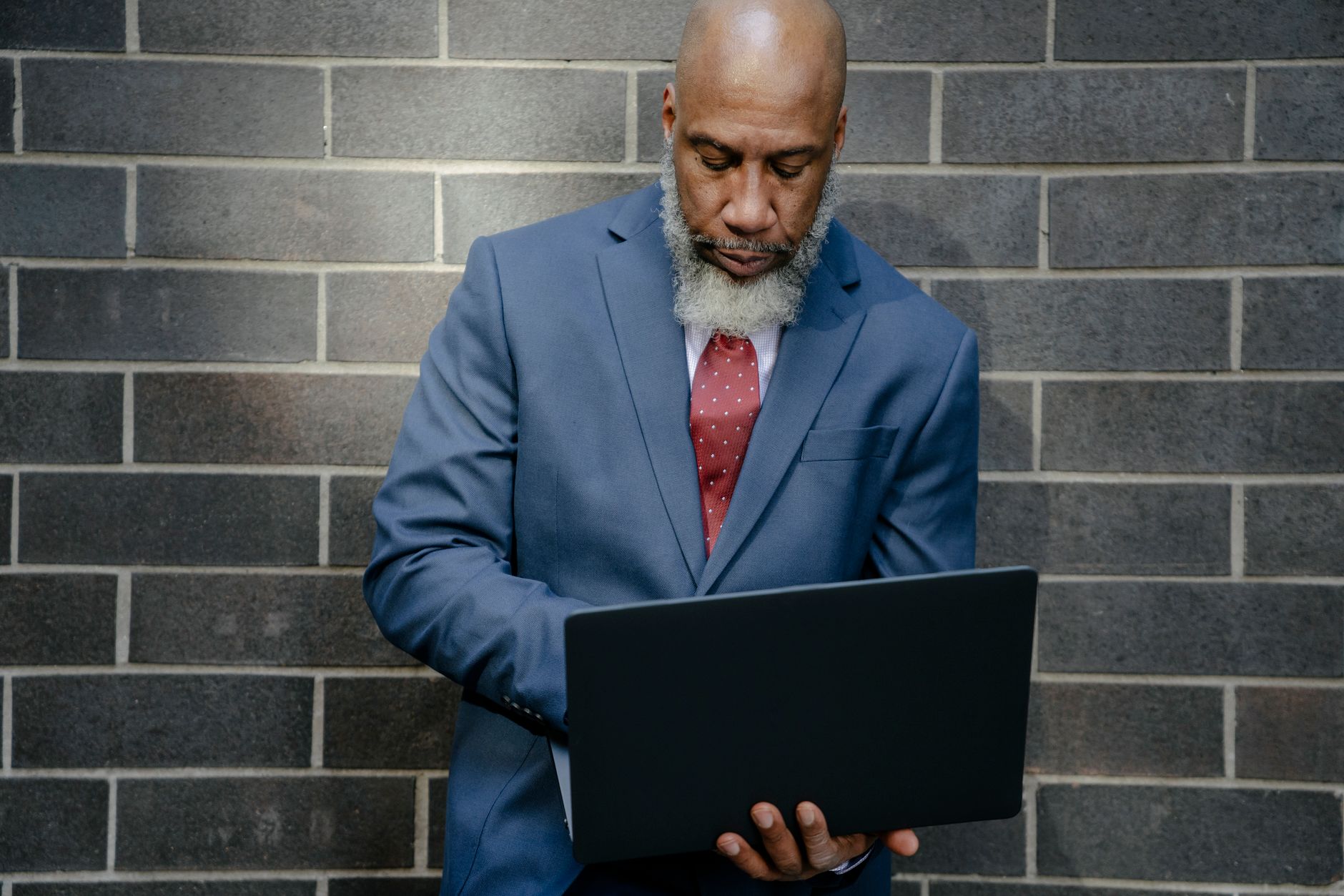 Bald African American man with a beard using a laptop against a brick wall, dressed in a blue suit.